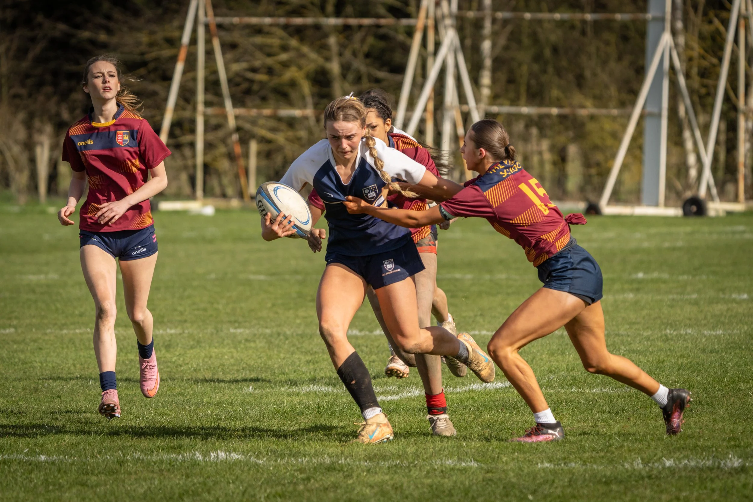 Girls playing rugby on a grassy field, with some trying to grab the ball from others, wearing sports jerseys and shorts.