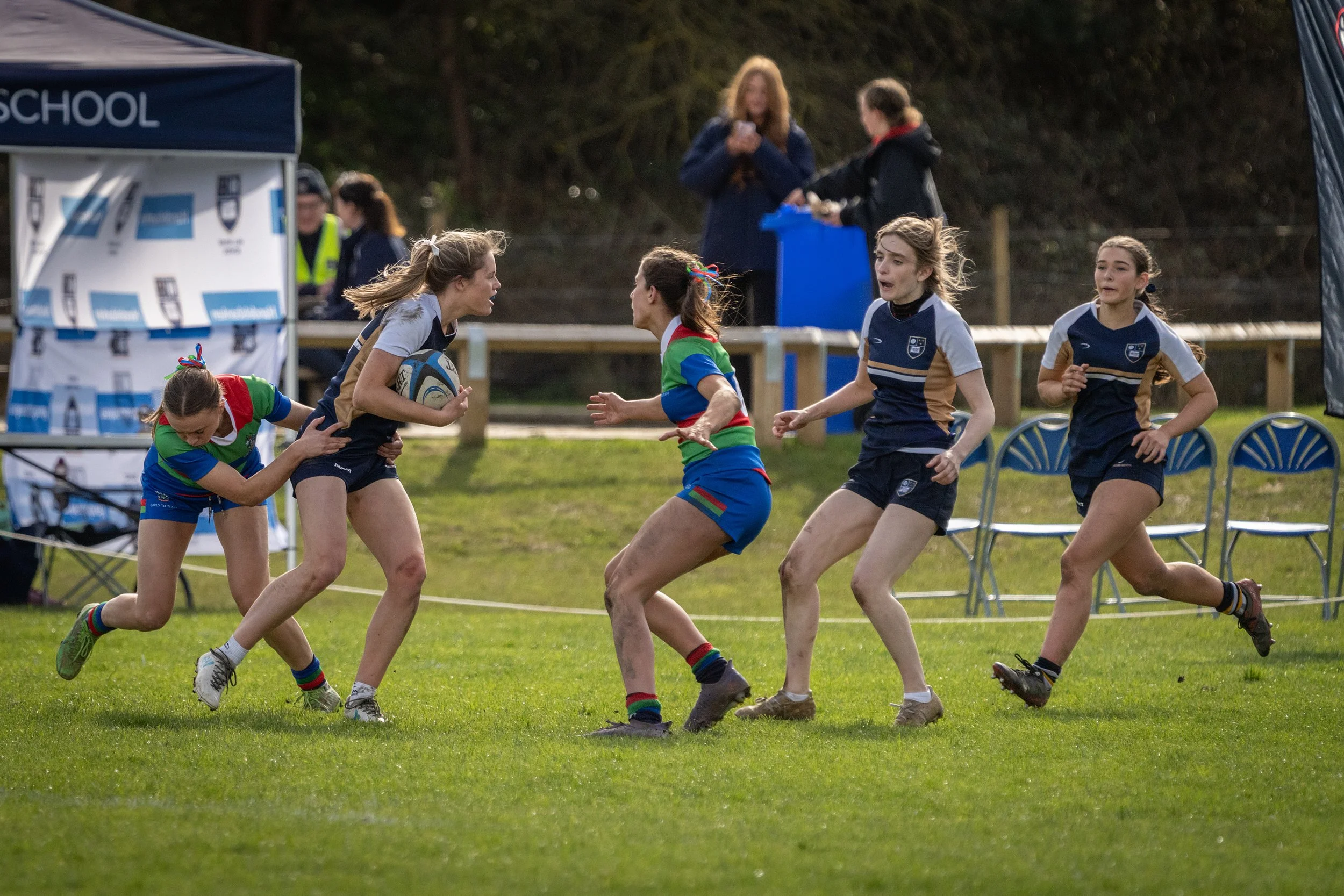Women playing rugby on a grassy field during a match, with spectators and tents in the background.