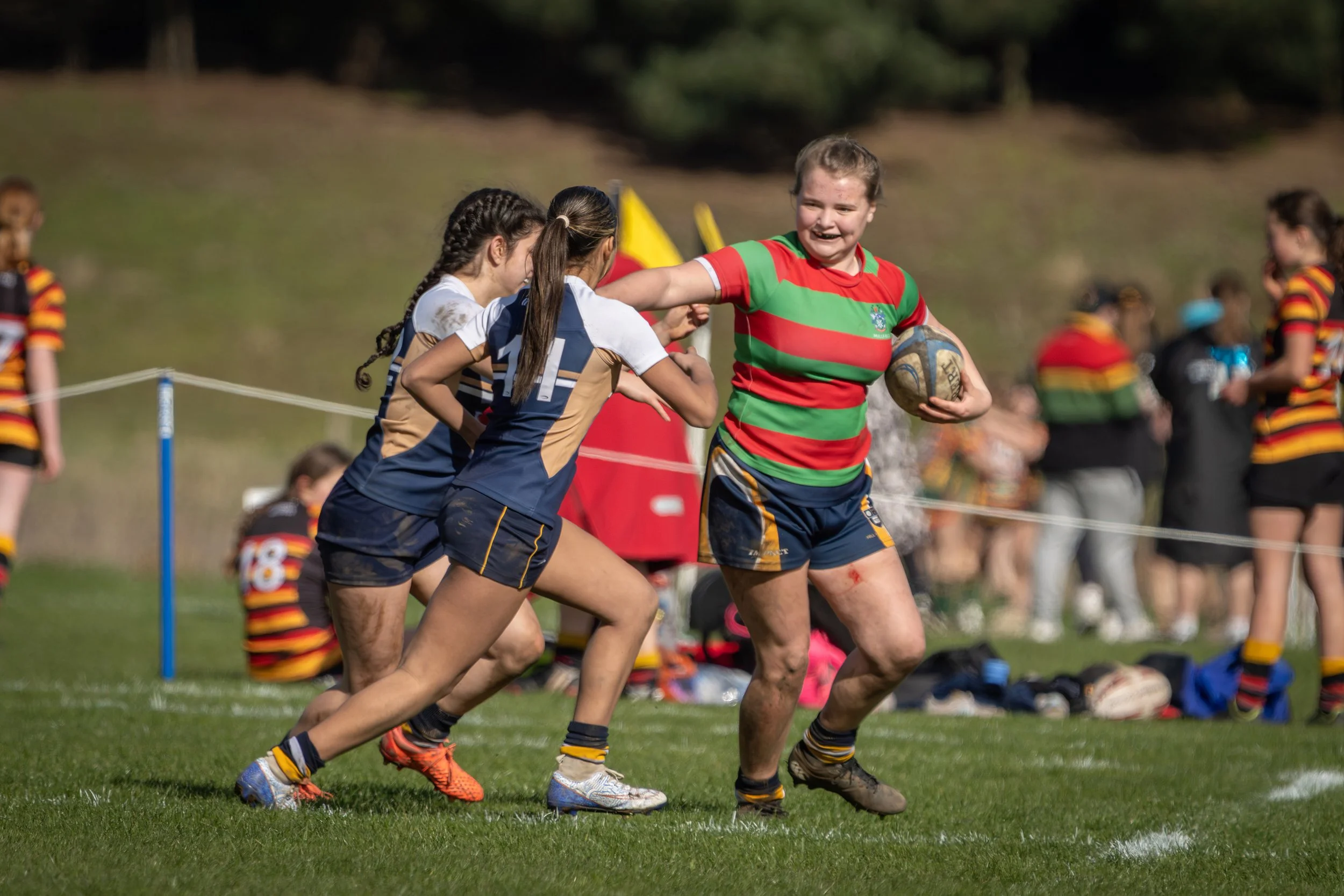 Young girls playing rugby on a grassy field, with one girl in a red and green striped jersey holding a rugby ball, engaged in a game against other girls in white and navy jerseys, with blurred spectators in the background.
