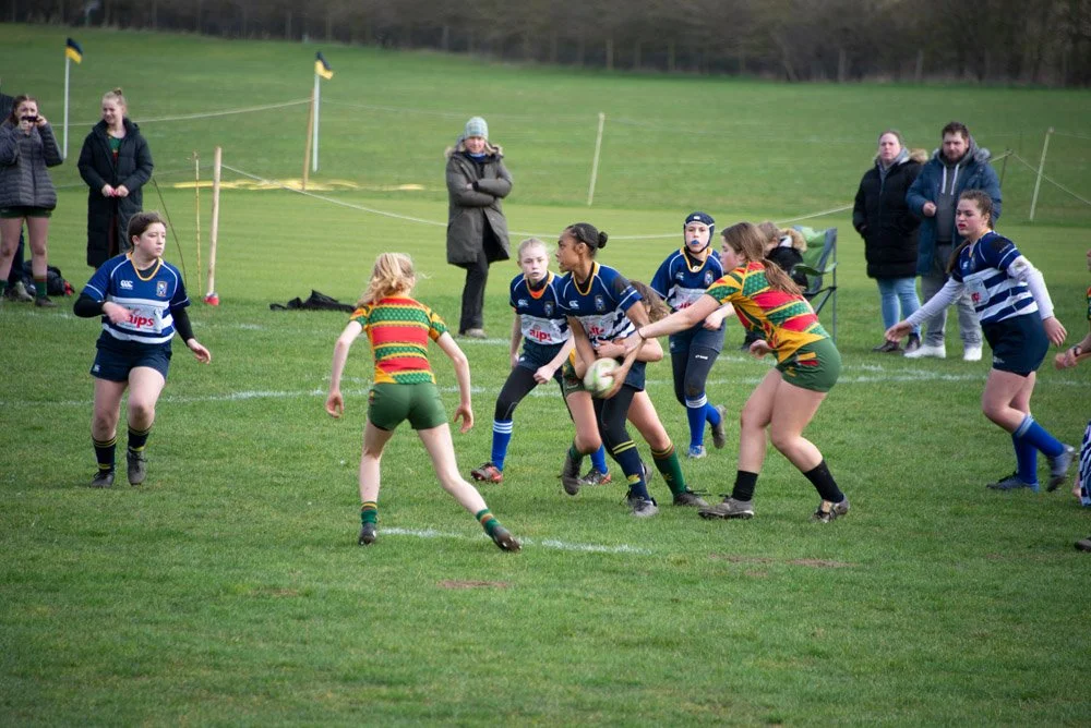 Girls playing rugby on a green field, some in striped jerseys, engaged in a game, with a few spectators nearby.