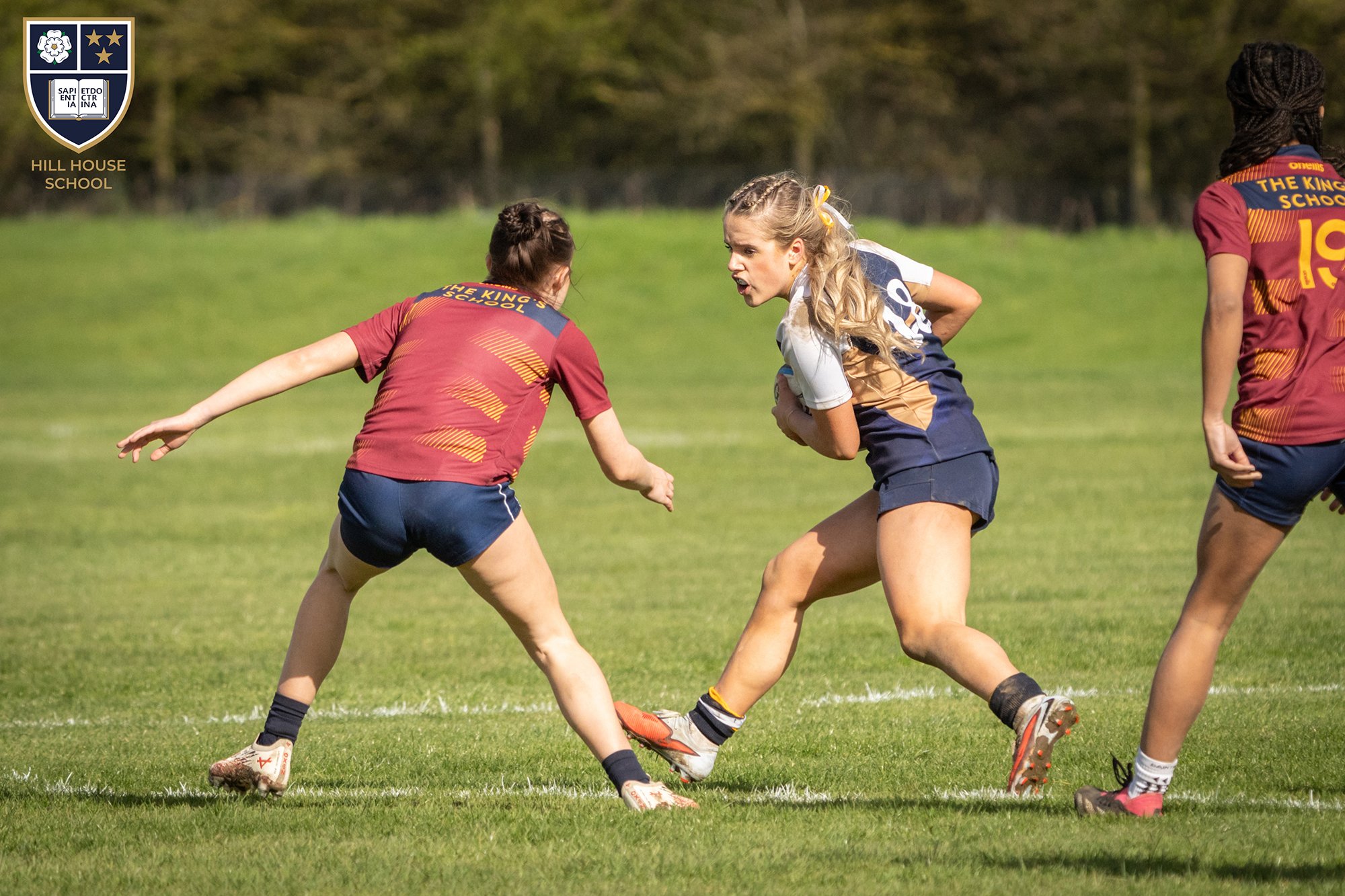 A group of young female rugby players in colorful uniforms engaging in a match on a grassy field. One girl appears to be holding a rugby ball while another is preparing to tackle or block. The scene takes place outdoors with trees in the background.