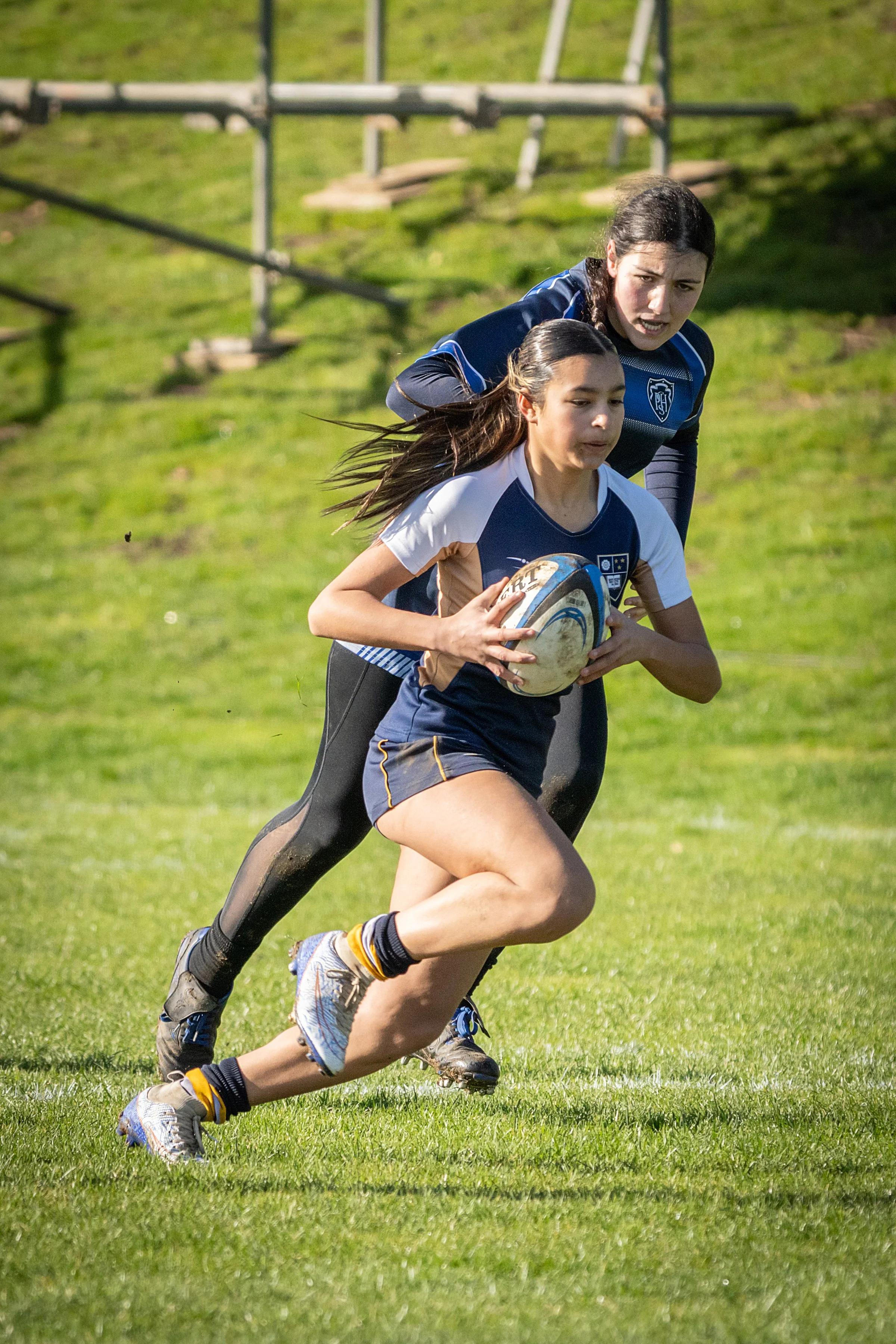 Two young women playing rugby on a grassy field, one preparing to pass the ball while the other runs alongside.