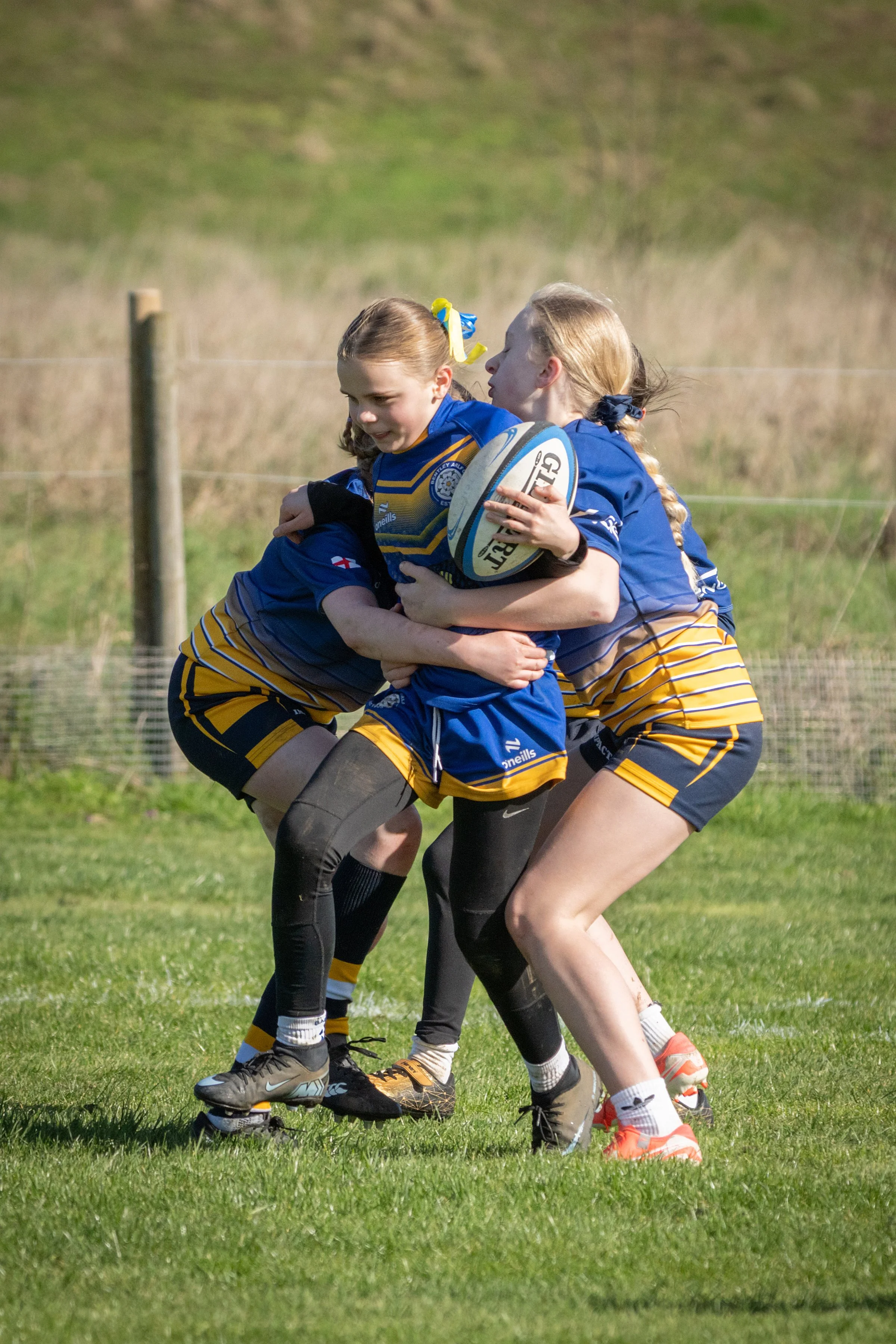 Two girls playing rugby on a grassy field, wearing blue and yellow uniforms, embracing each other during the game.