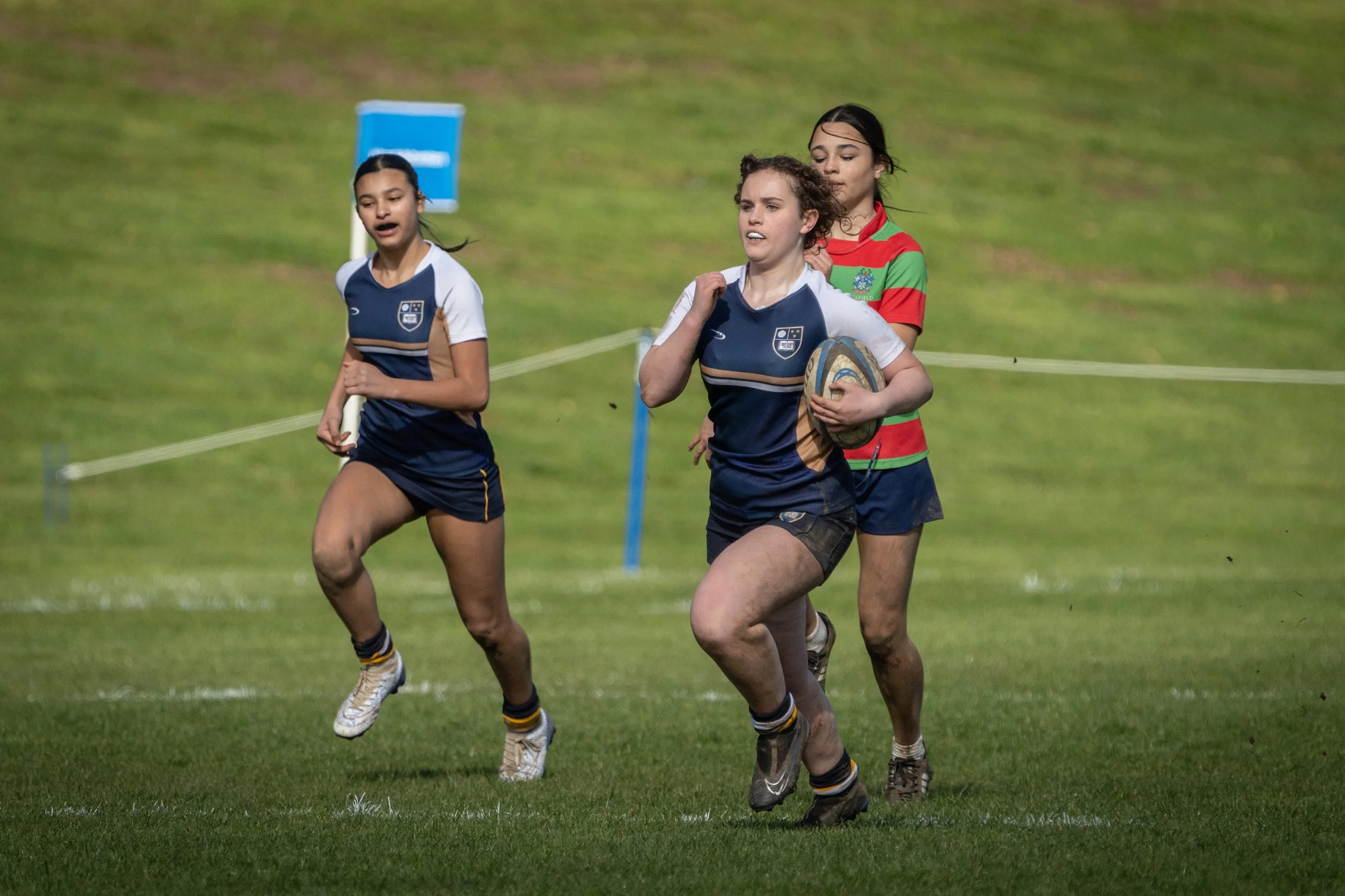 Young girls playing rugby on a grassy field, with one girl holding a rugby ball and wearing a navy and white uniform, closely followed by two other teammates.