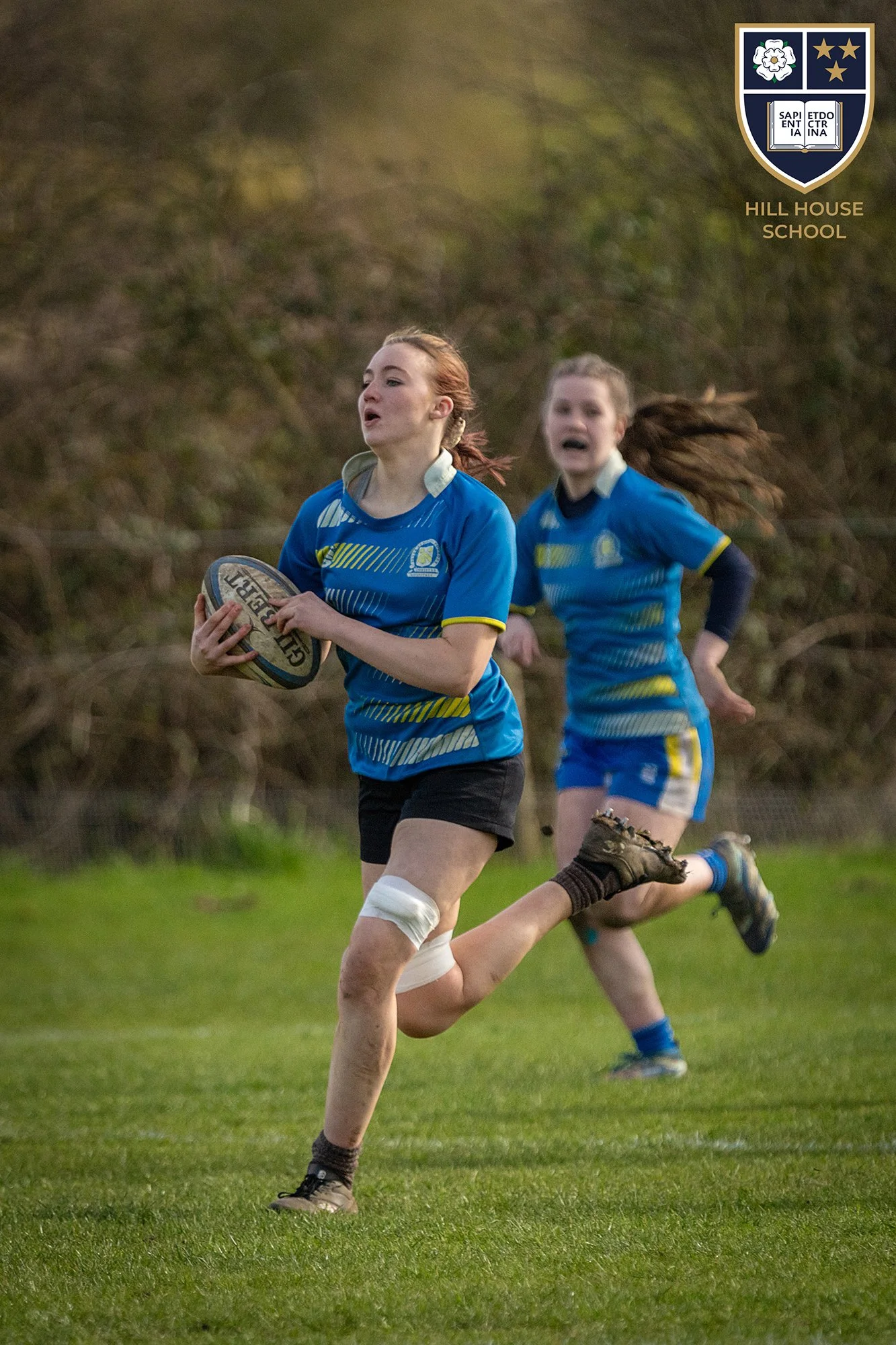 Two young girls playing rugby on a grassy field, wearing blue sports uniforms, with the Hill House School crest in the top right corner.