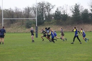 A rugby game on a grassy field with players running and trying to score as the goalkeeper watches.