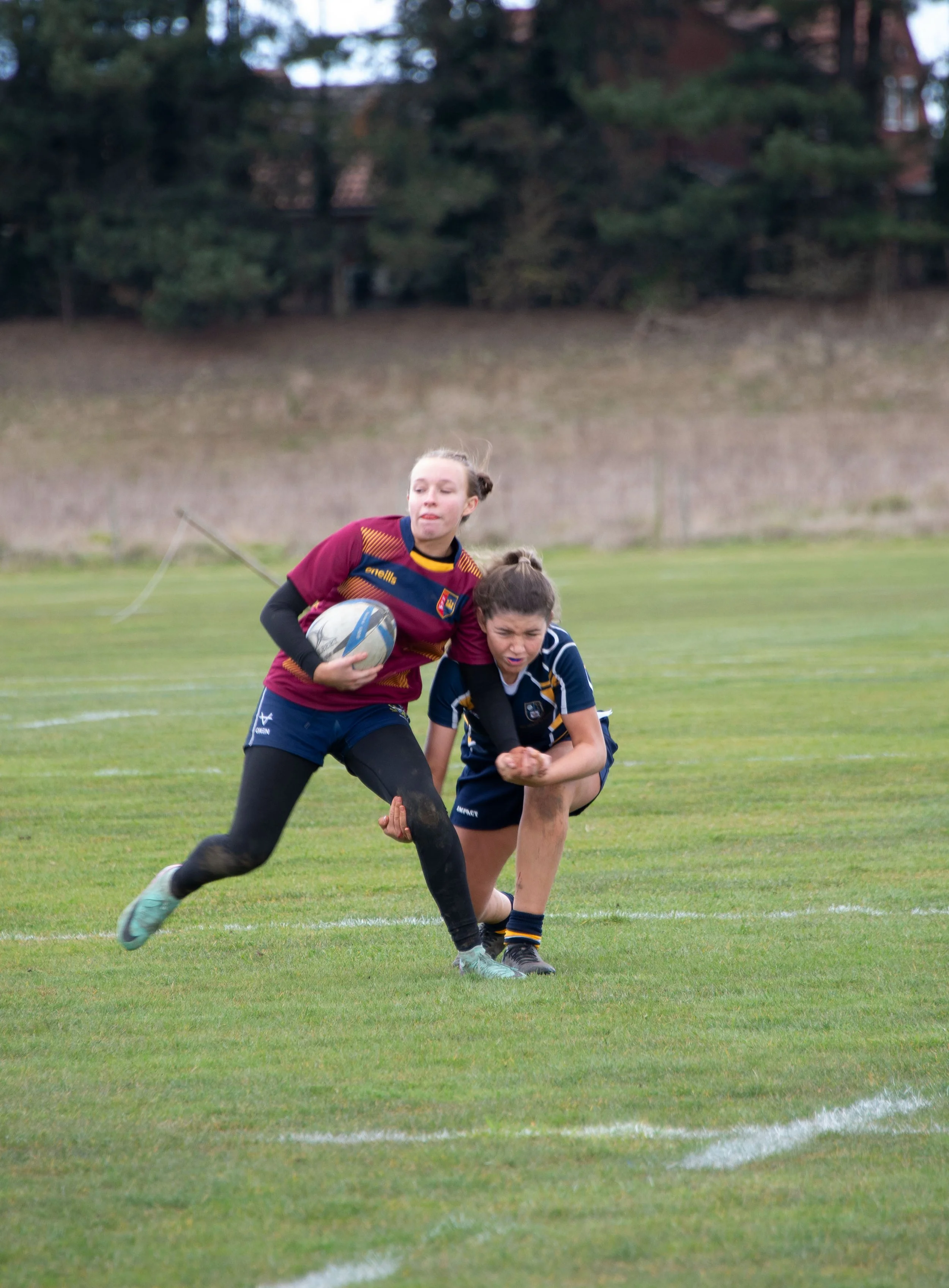 Two young girls playing rugby on a grass field, one in a maroon and blue uniform holding a rugby ball, the other in a dark blue and yellow uniform, both engaged in a tackle.