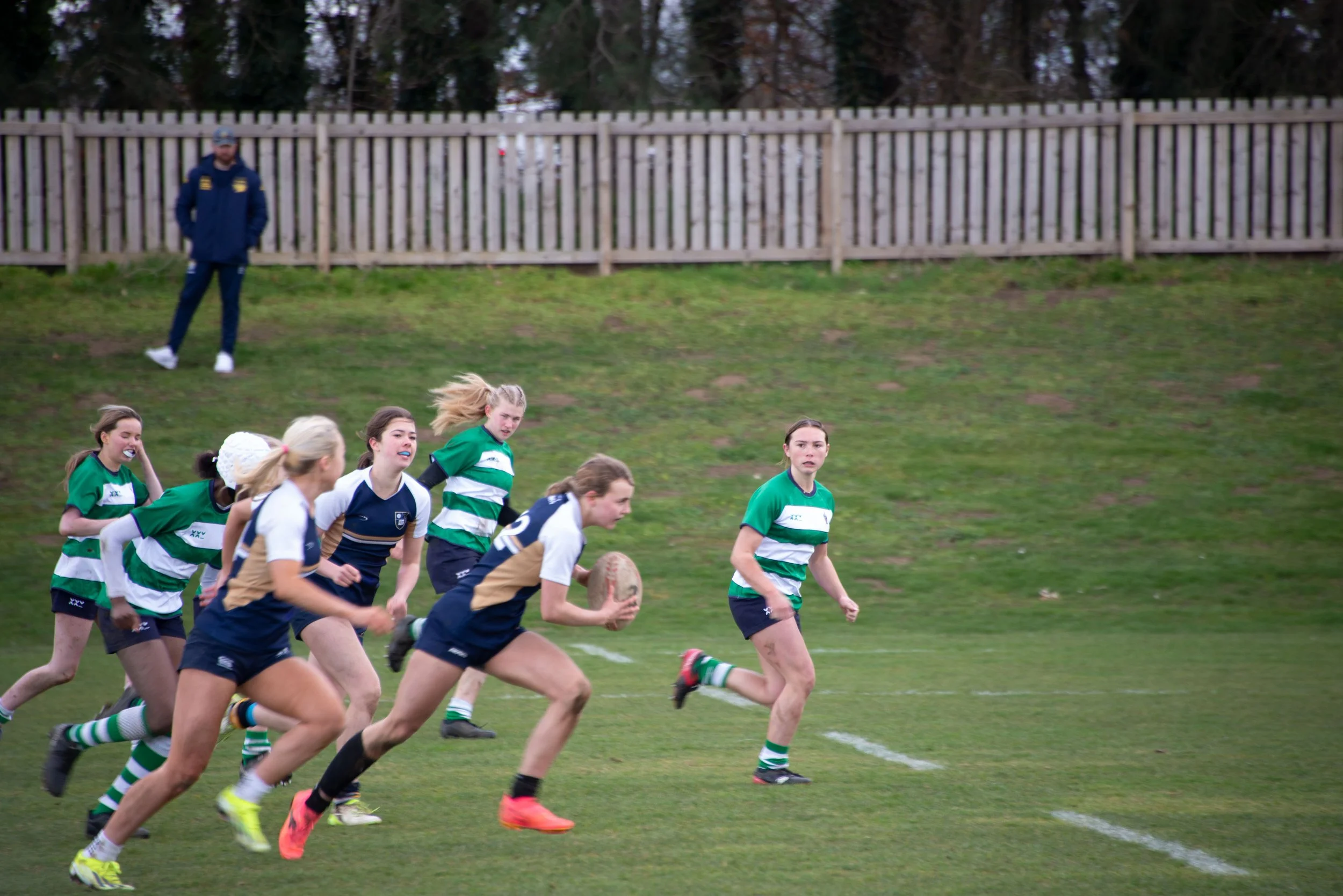 Women playing rugby on a grassy field with a wooden fence and trees in the background