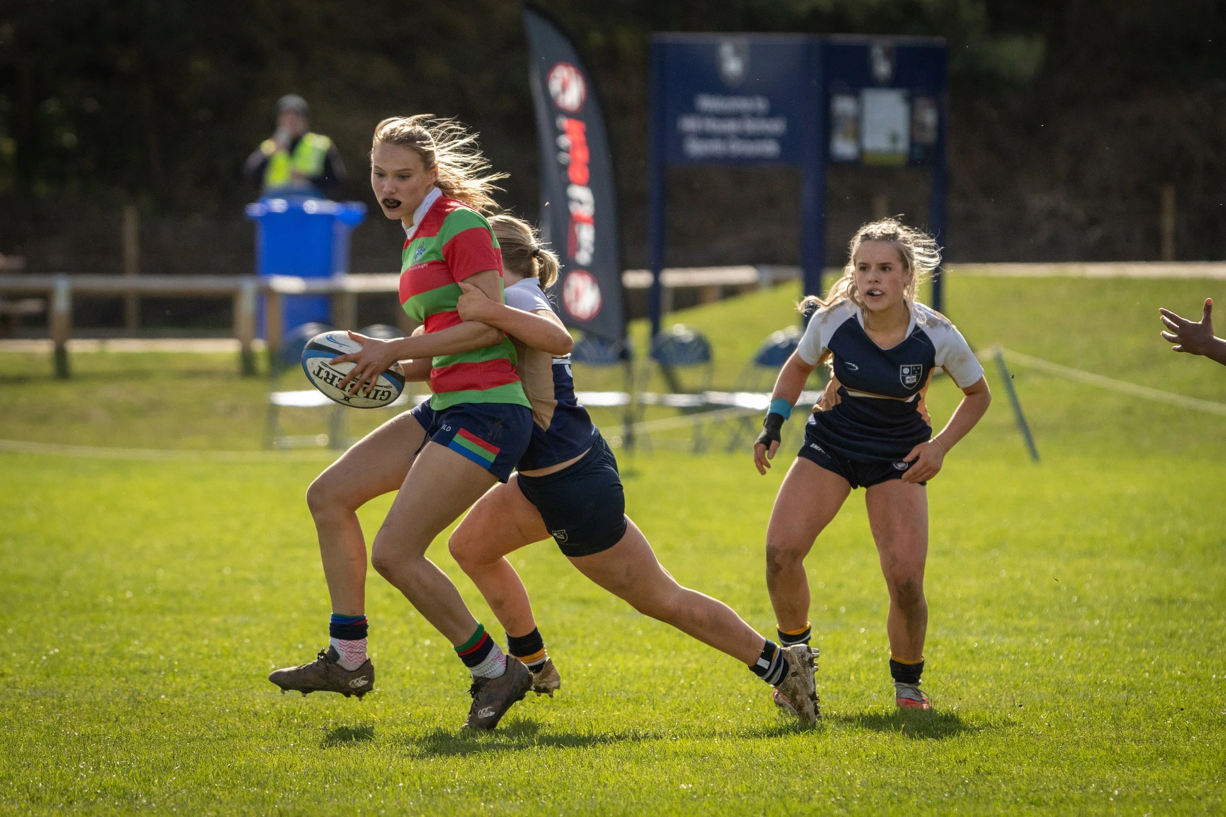 Two young women playing rugby on a grassy field during daytime with a third person in the background. One woman is wearing a striped red and green jersey, holding a rugby ball, and running while being tackled by another woman in a dark blue and white