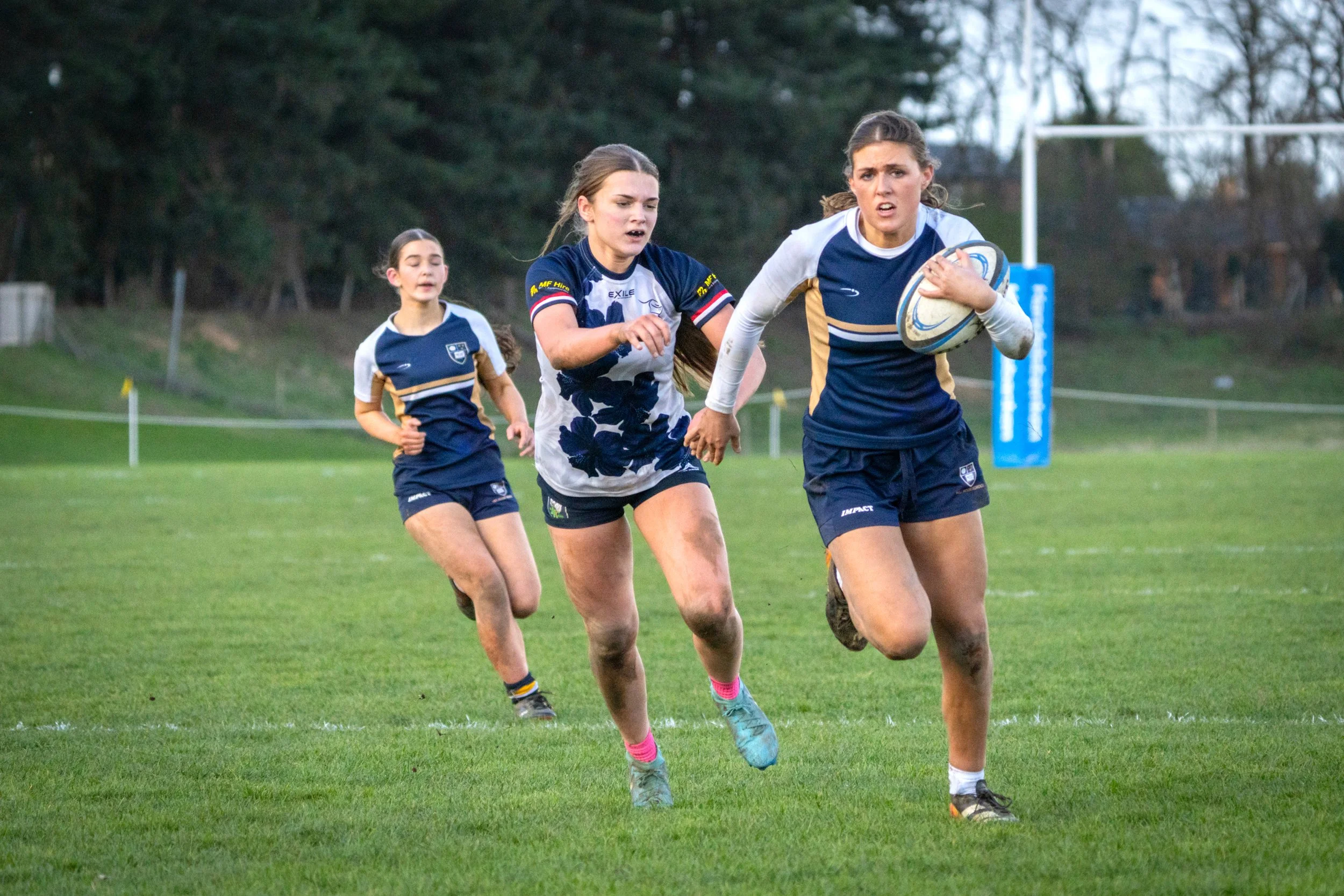 Three young women playing rugby on a grassy field, with trees in the background. The woman in the foreground is running with a rugby ball, wearing a navy and white jersey with pink socks. The other two women are running behind her, one wearing a simi