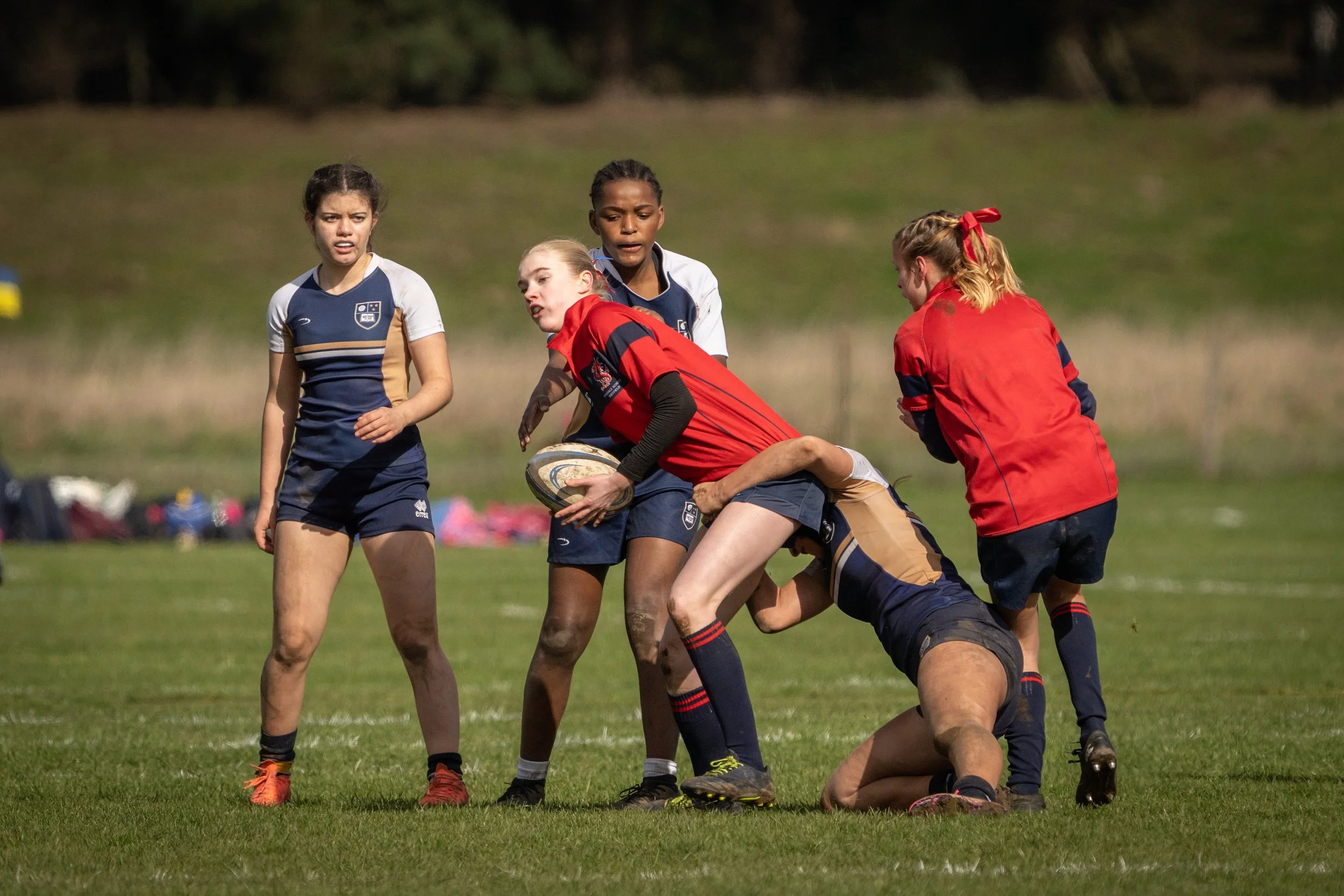 Women playing rugby on a grassy field, with one woman holding the rugby ball while others attempt to block or take her down during daytime.