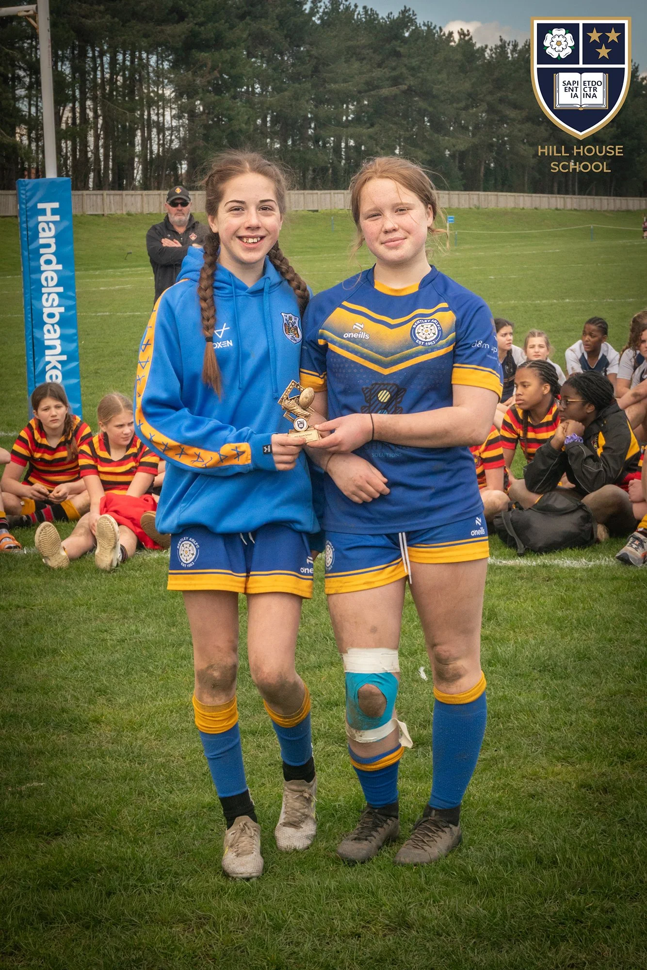 Two young female rugby players in blue and yellow uniforms holding a small trophy, standing on a grassy field with other players sitting behind them, and a man in sunglasses standing in the background. A crest with a white flower, stars, and an open 