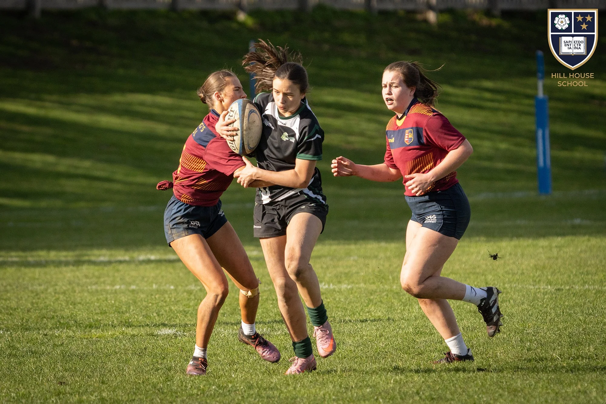 Three female rugby players in red and black uniforms competing for the ball during a game on a grass field, with a Hill House School logo in the top right corner.