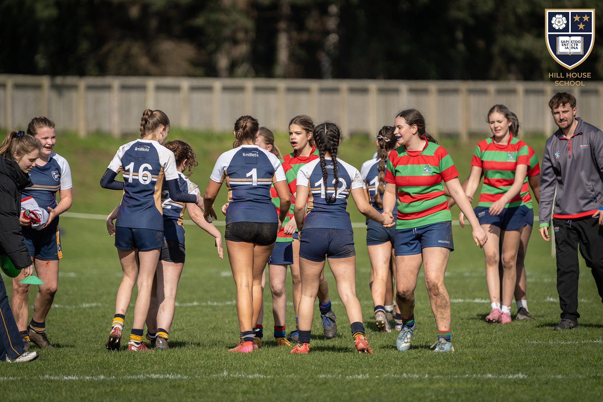 A group of girls in sports uniforms and a coach on a grassy field after a rugby game, some holding hands, with a fence and trees in the background.