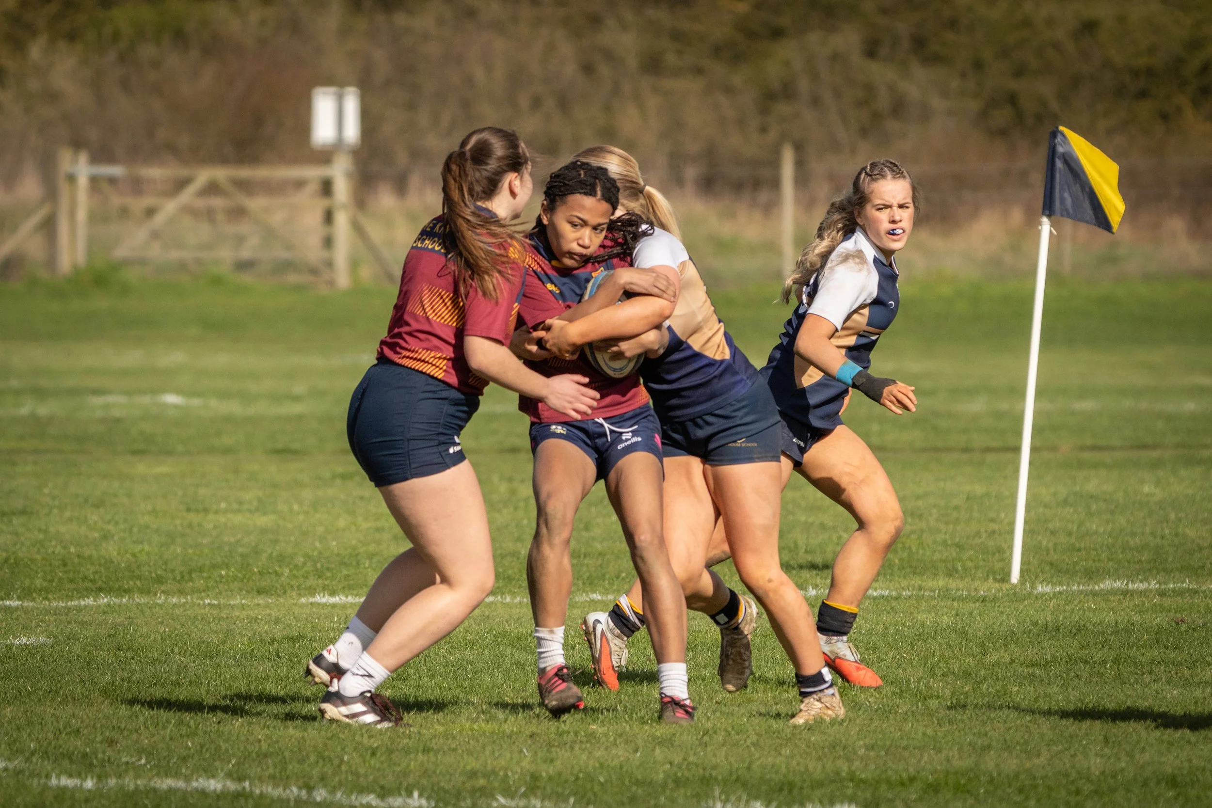 Young women playing rugby on a grassy field, attempting to gain control of the ball near a corner flag.