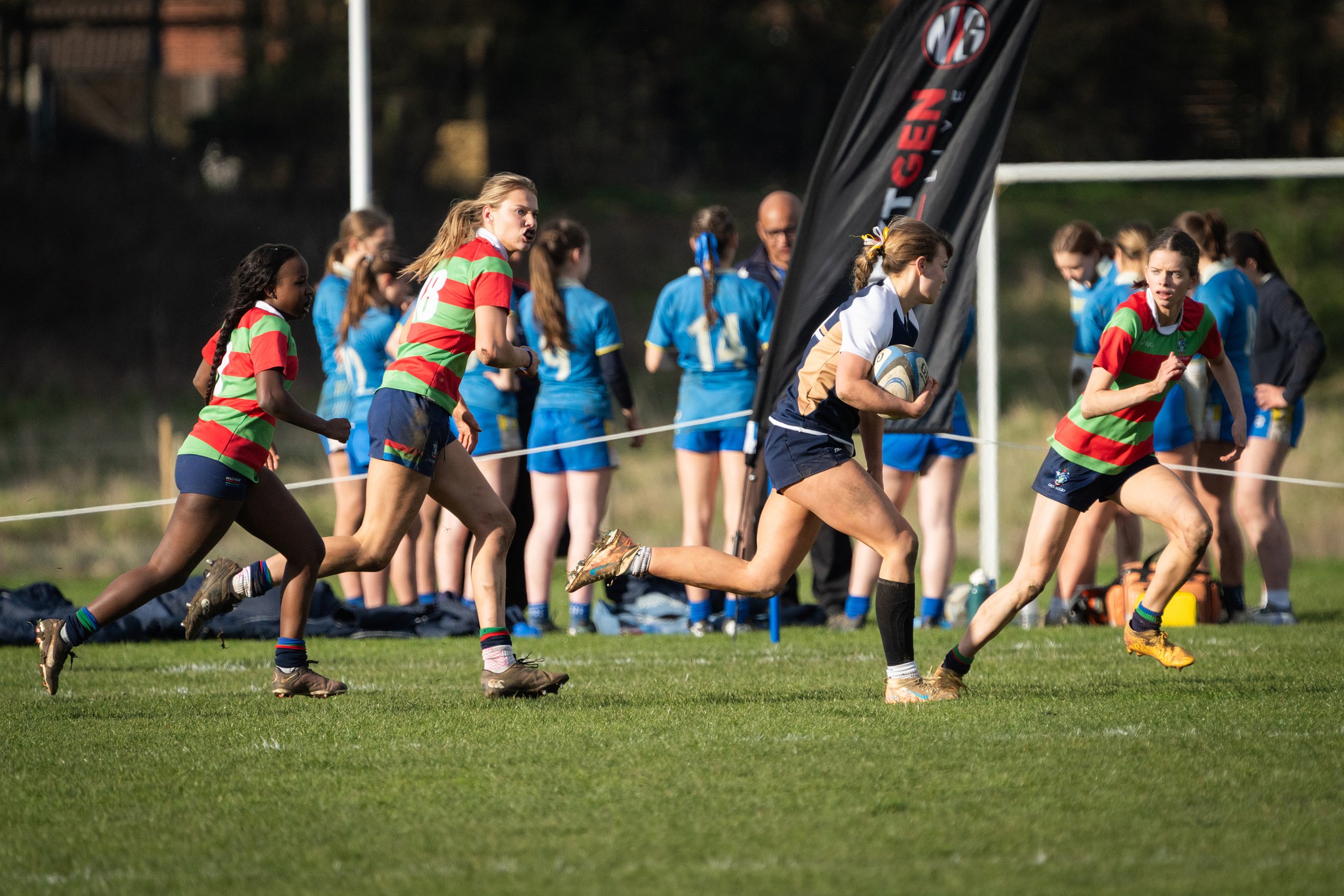 Women's rugby game with players running on field, some holding rugby ball, in athletic uniforms, with spectators or coaches in background.