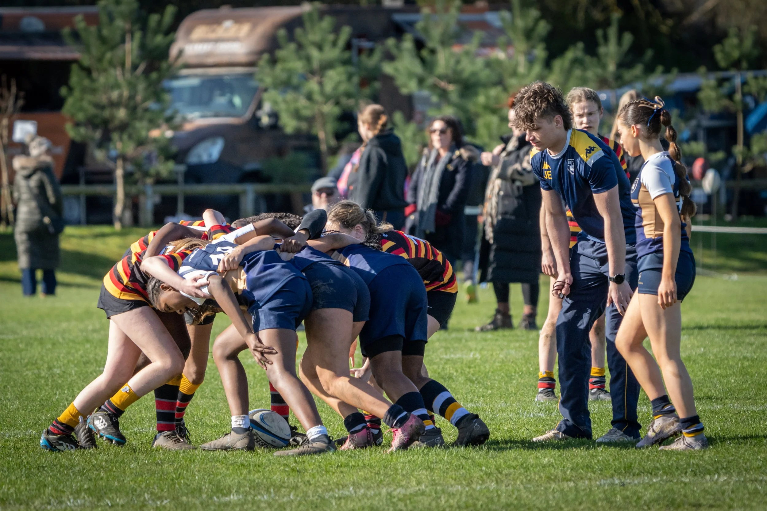 A group of female rugby players in a scrum on a grassy field, with a coach or referee observing. In the background, spectators and parked cars are visible.