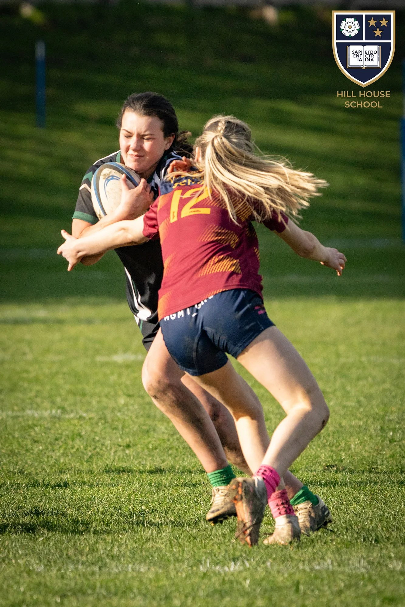 Two women playing rugby on a grassy field, one in a black and green uniform holding a ball, the other in a maroon and yellow uniform attempting to tackle.