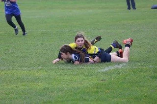 Two young women playing rugby on a grassy field, tackling each other on the ground during a game.