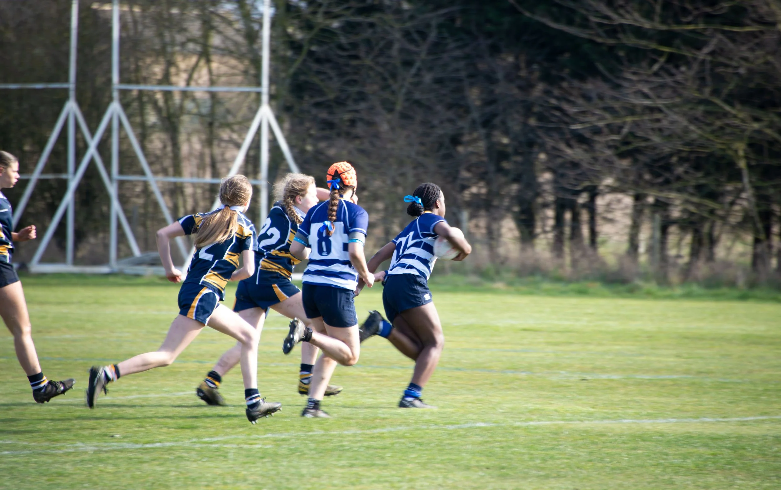 Girls rugby players in blue and yellow striped uniforms running on a field during a match.