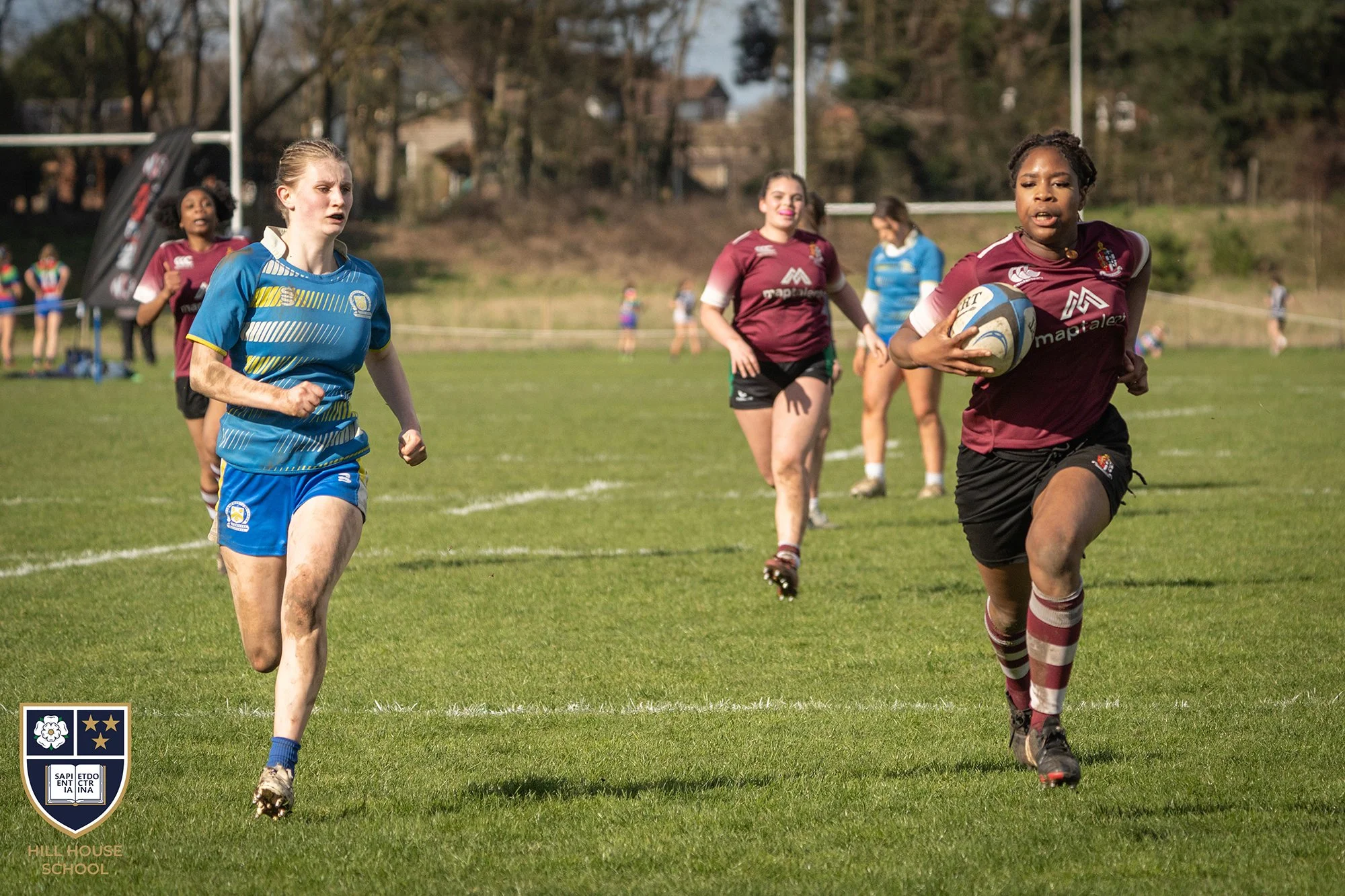 Girls playing rugby on a grassy field during daytime. One girl in maroon and white uniform is holding a rugby ball, running while others are chasing her. Trees in the background, some people watching along the side, and a school emblem in the bottom 