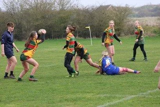 Girls playing rugby on a field, tackling a girl in blue uniform, with two other girls and a boy nearby.