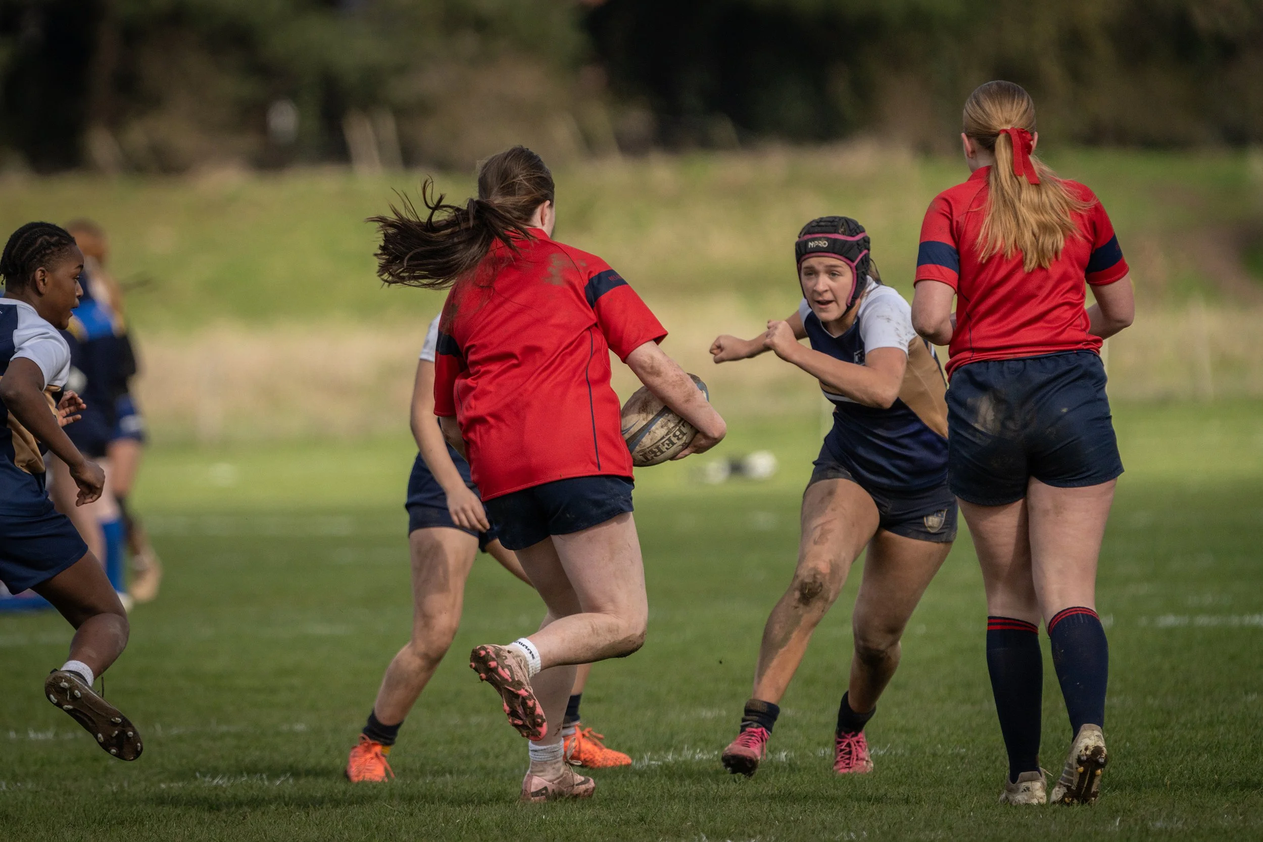 Group of young women playing rugby on a grassy field, some with muddy uniforms and a rugby ball in play.