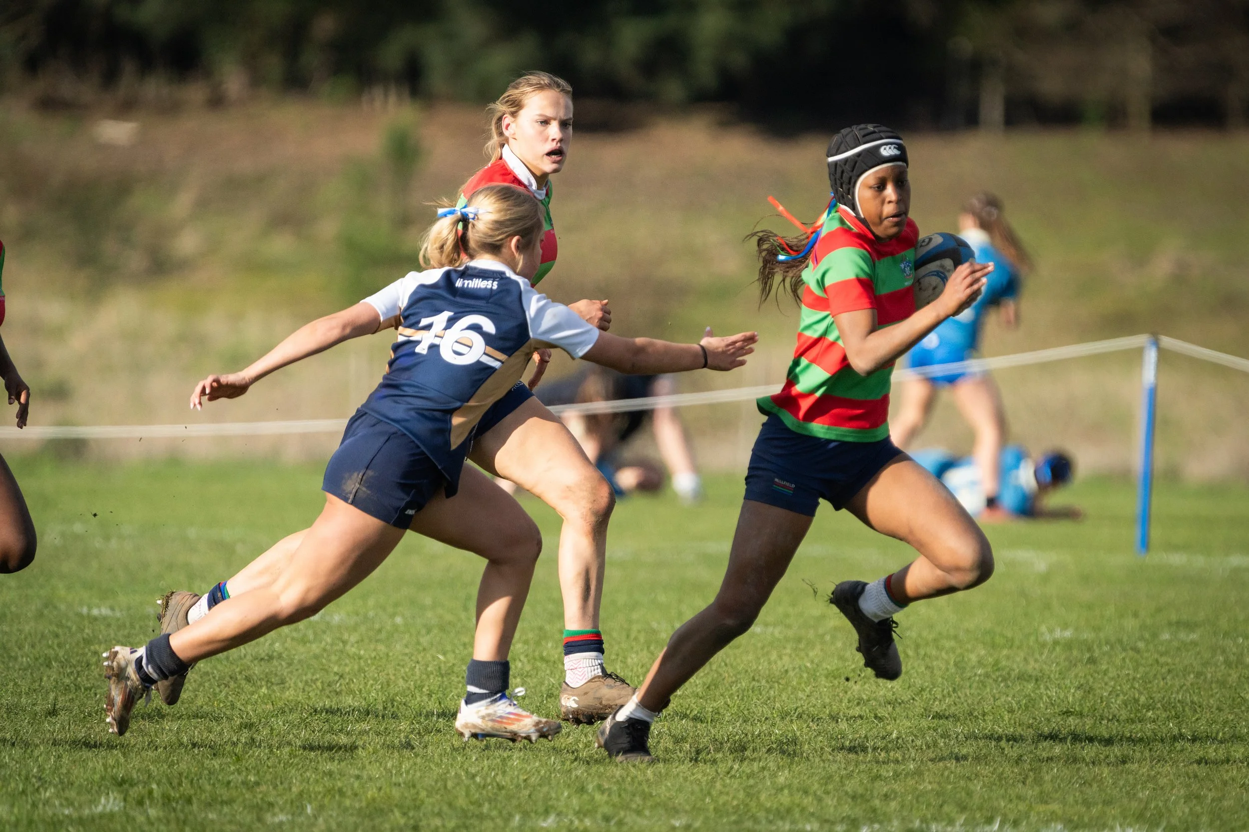 Group of young girls playing rugby on a grassy field during daytime. One girl in a striped red and green shirt is holding a rugby ball, while others in sport uniforms chase her.