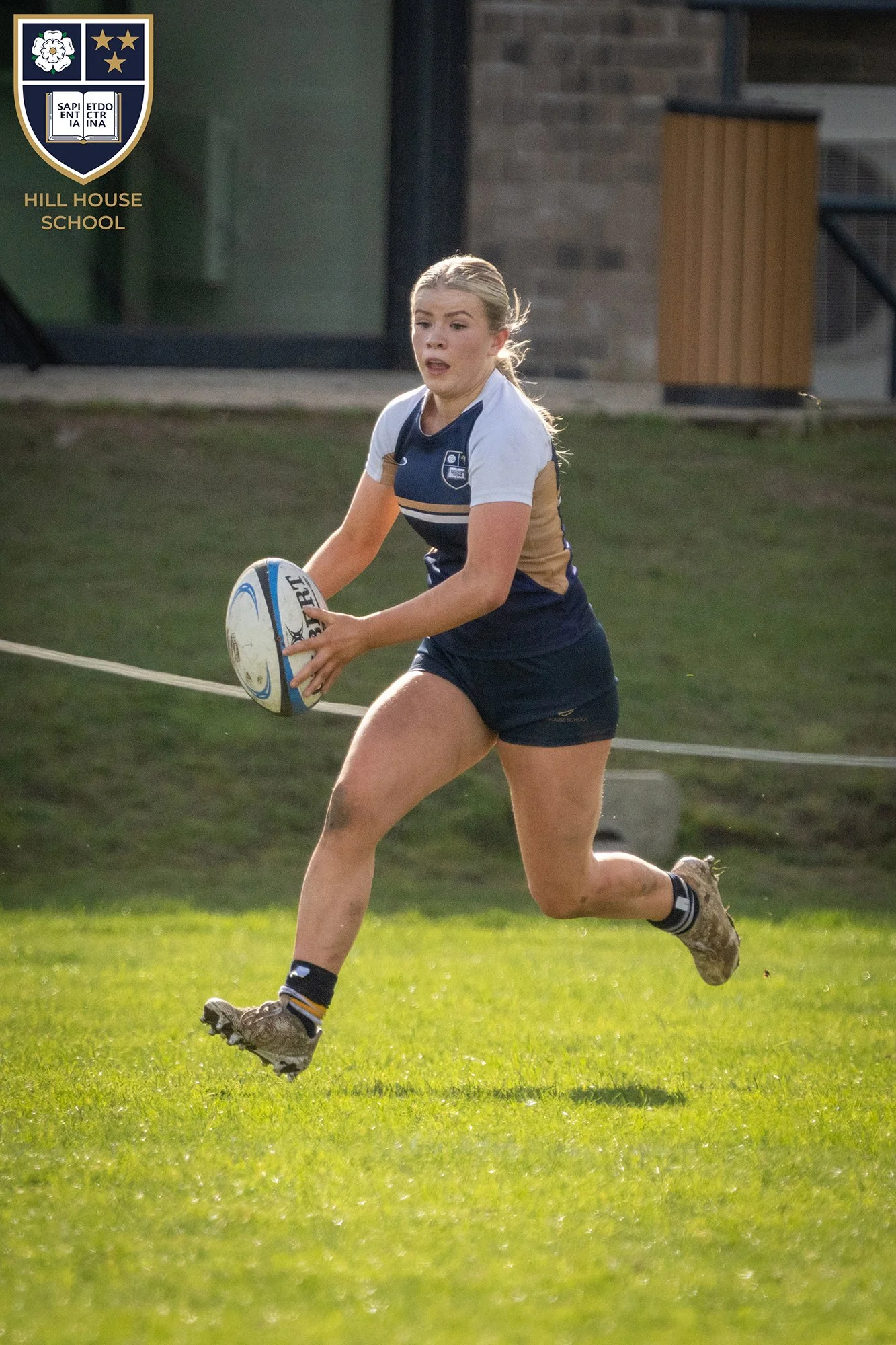 A female rugby player in a navy and white jersey running across a grass field while holding a rugby ball, with a building in the background.