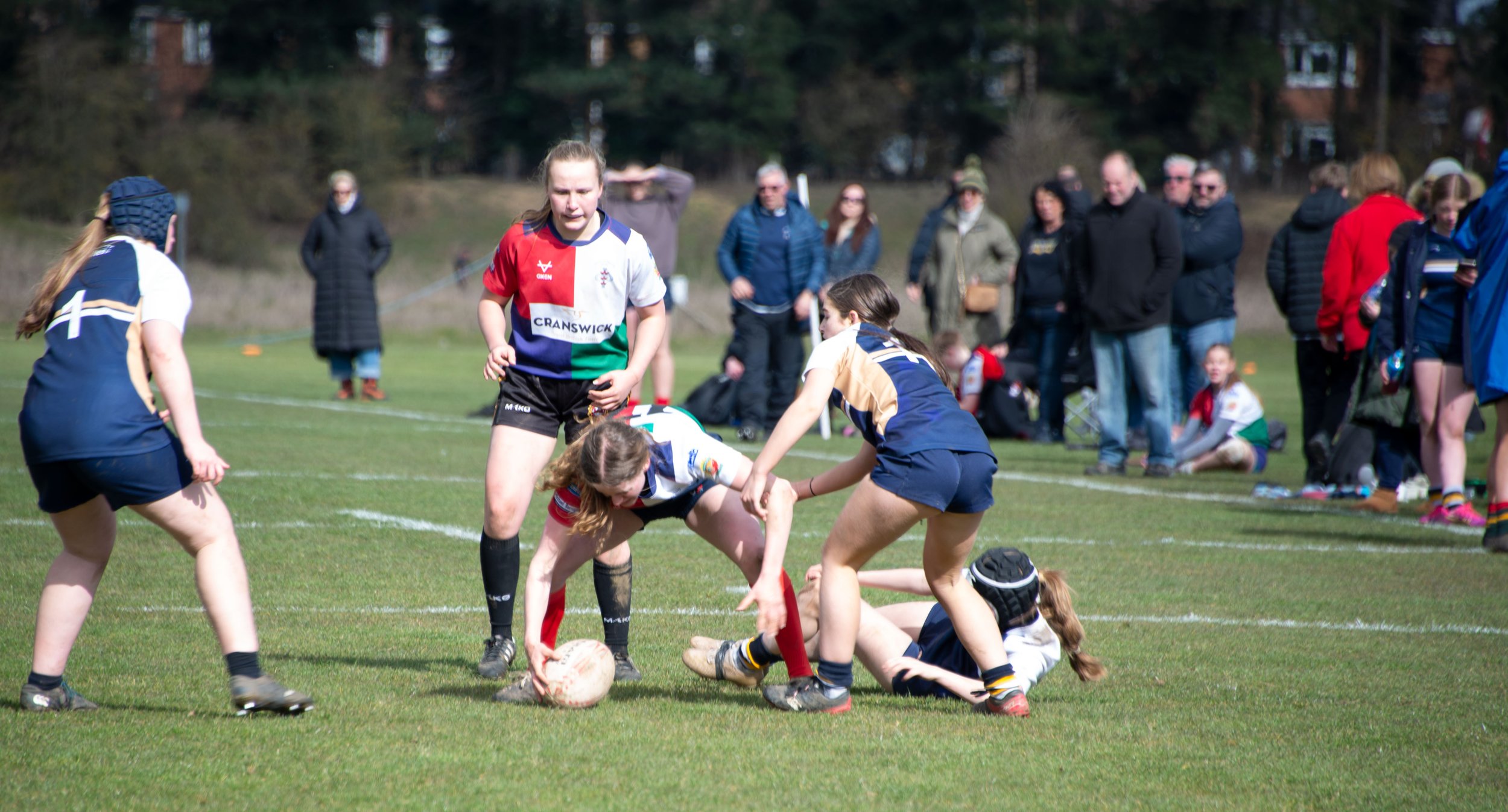 Women's rugby match with players tackling and holding the ball on a grassy field, spectators watching in the background.