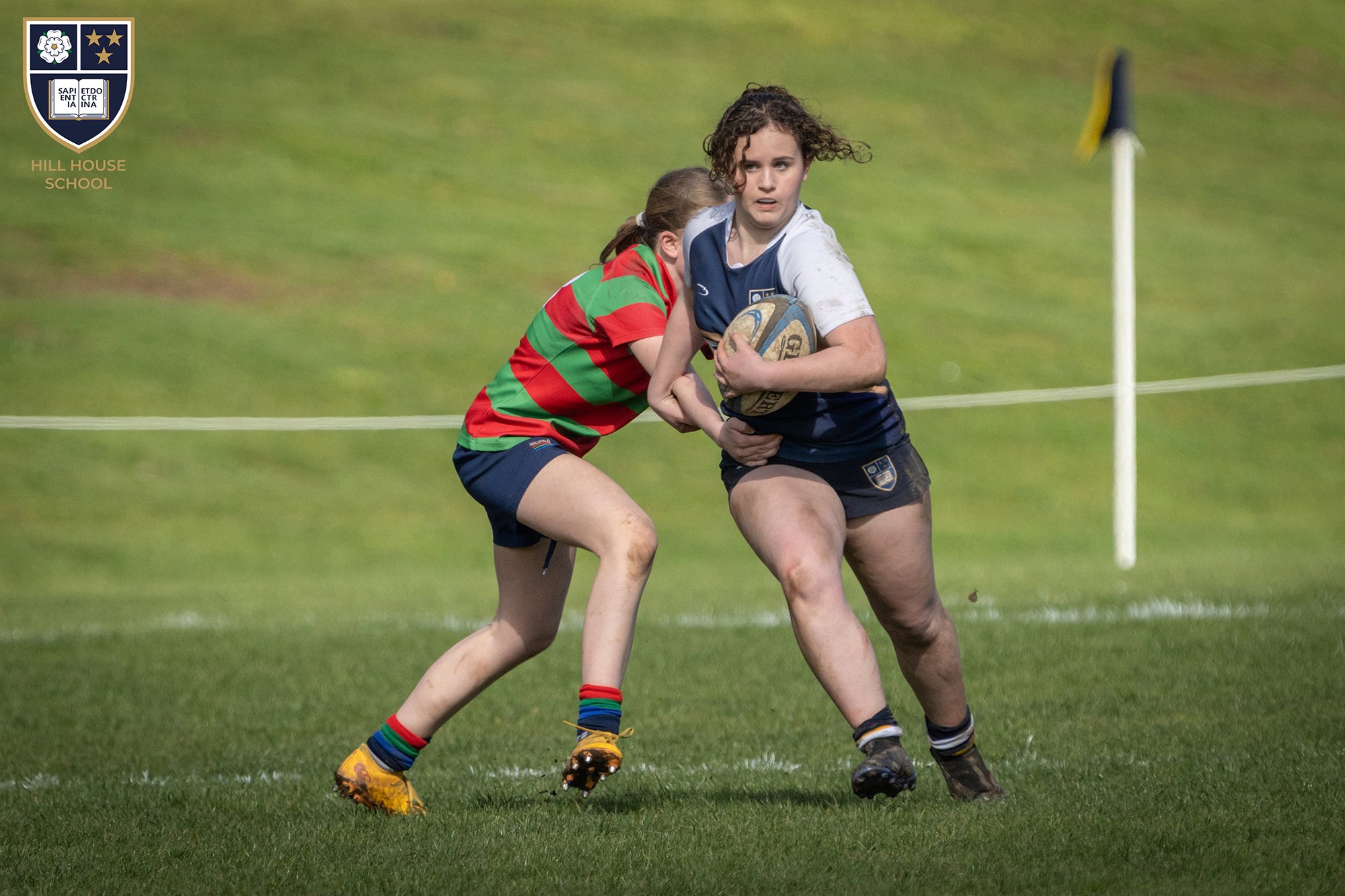 A rugby game featuring two female players, one in a white and blue uniform running with ball and another in a red and green striped uniform attempting to stop her, on a grass field during night time.