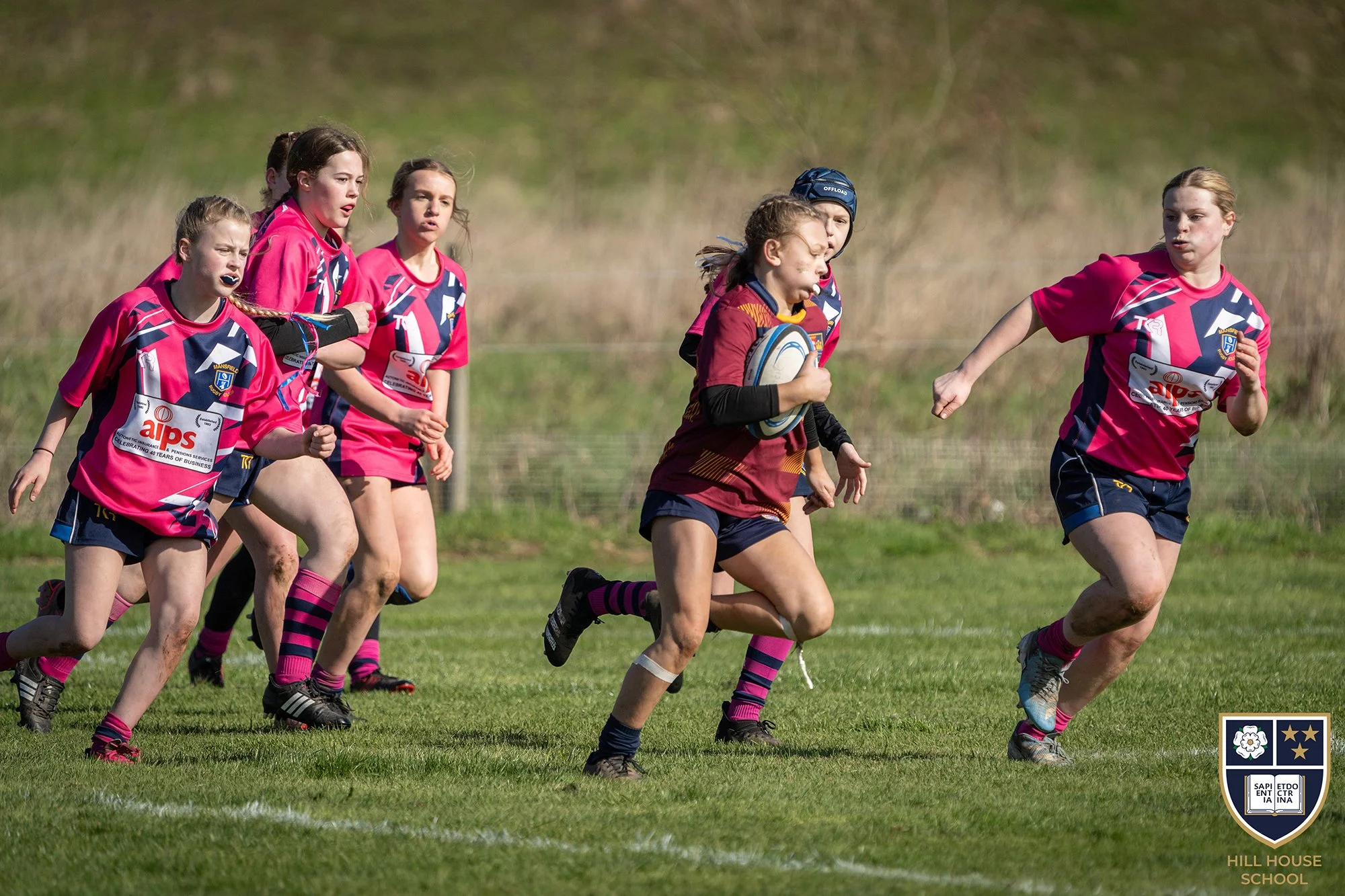 Young girls playing rugby on a grassy field during daytime, wearing pink and maroon jerseys, with one girl holding a rugby ball, indicating a rugby match among girls from Hill House School.
