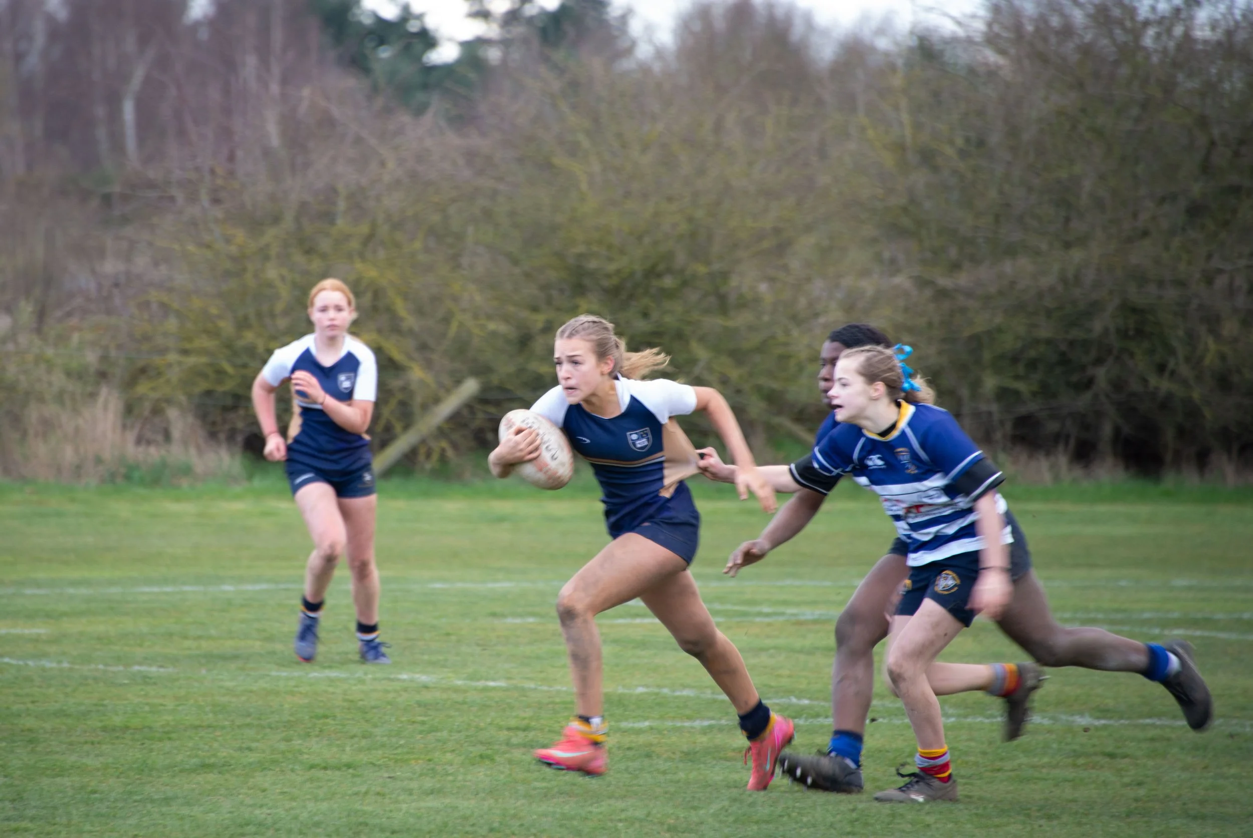 Girls playing rugby on a grassy field with trees in the background.