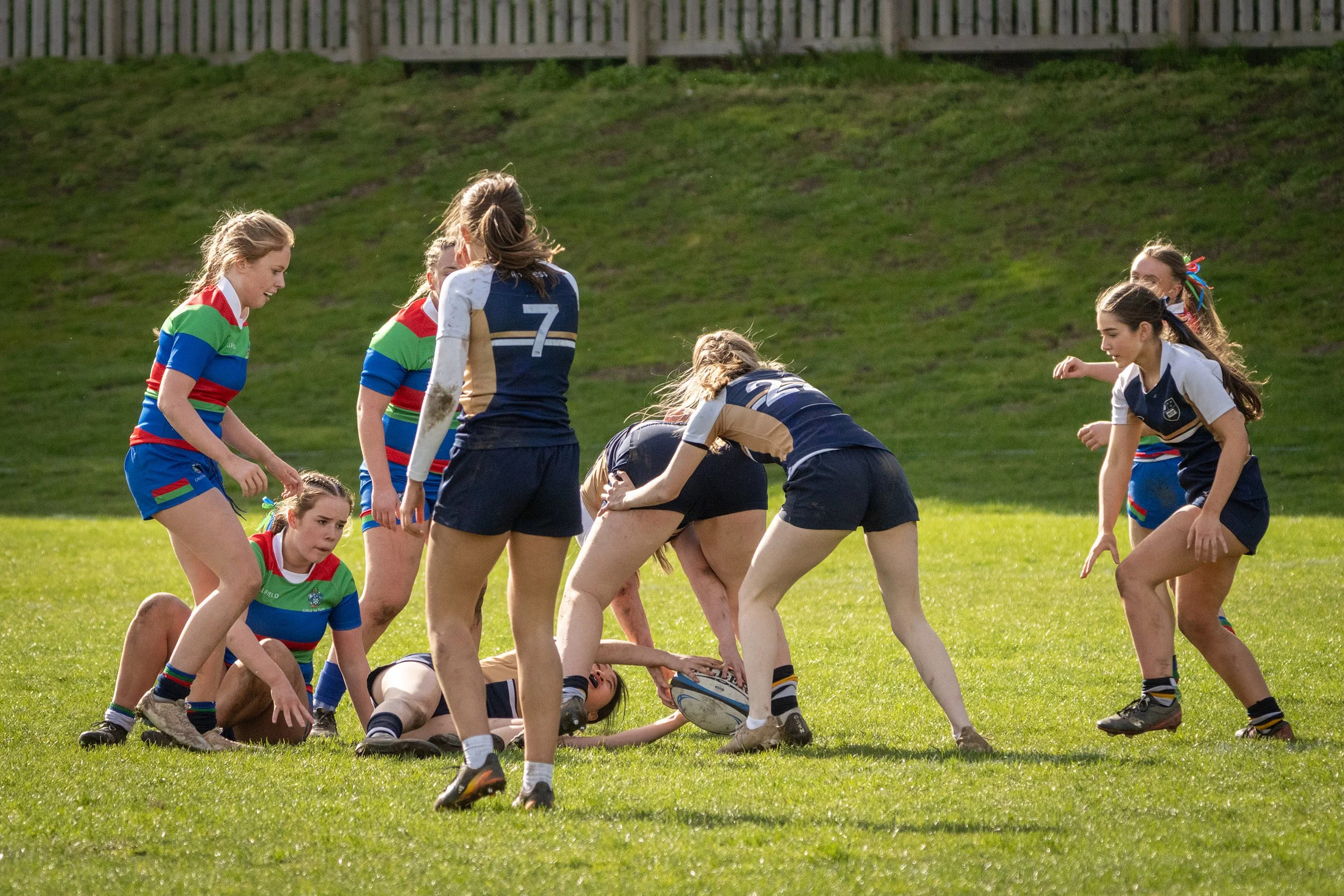 Women rugby players in colorful and navy uniforms competing on a grassy field, with one girl on the ground and others standing around her during daytime.
