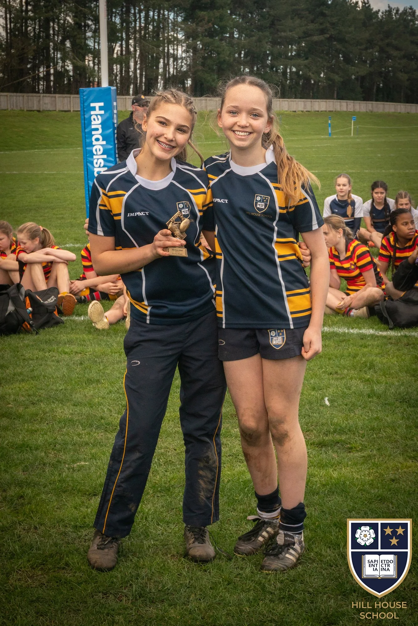 Two young female athletes in rugby uniforms standing on a grassy field smiling at the camera, with a group of other girls sitting behind them; one girl is holding a small award or trophy. The scene appears to be after a match or sports event at Hill 