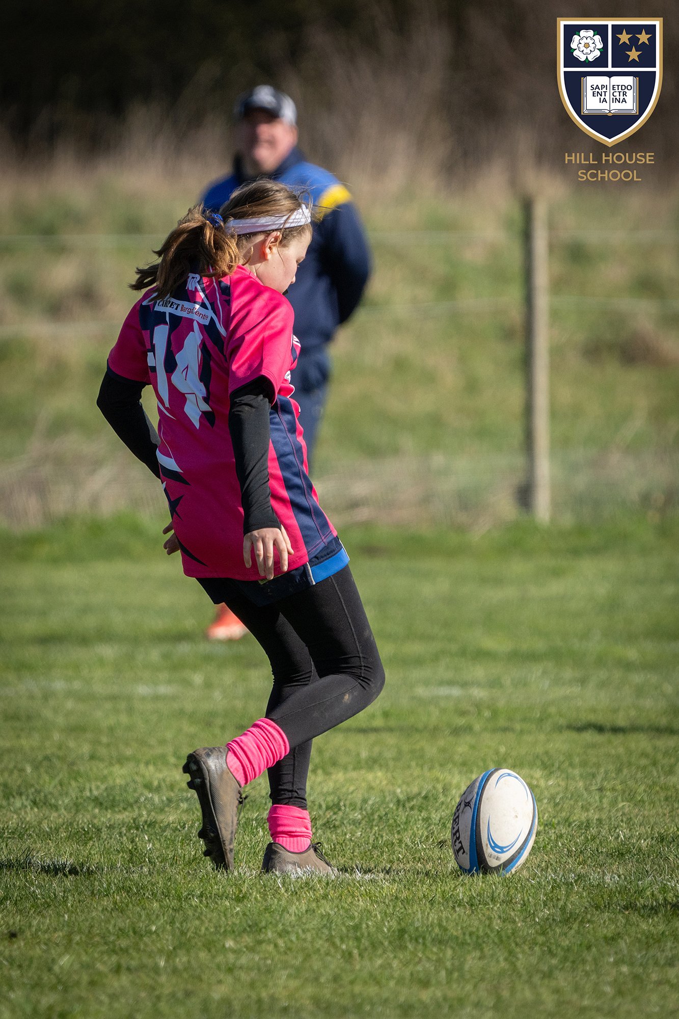 Young girl in a pink and black sports uniform kicking a rugby ball on a grassy field, with a coach watching in the background, in front of a hill and trees, with a Hill House School crest logo in the top right corner.