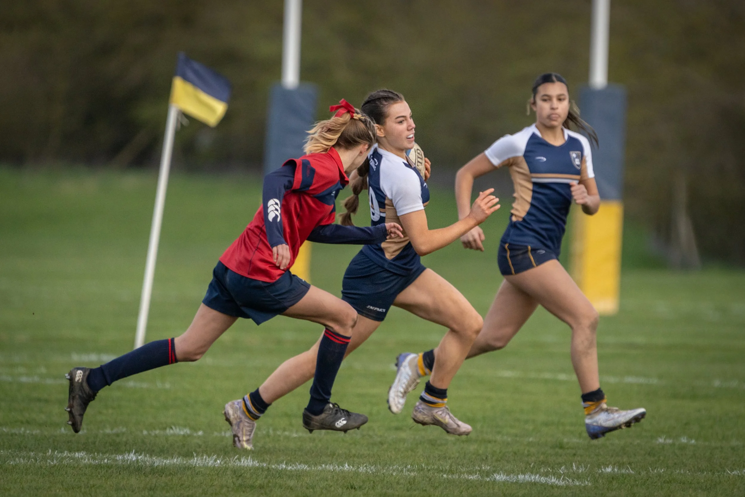 Women playing rugby on a grassy field, wearing blue and gold jerseys, and running with a rugby ball. There is a Ukrainian flag in the background.