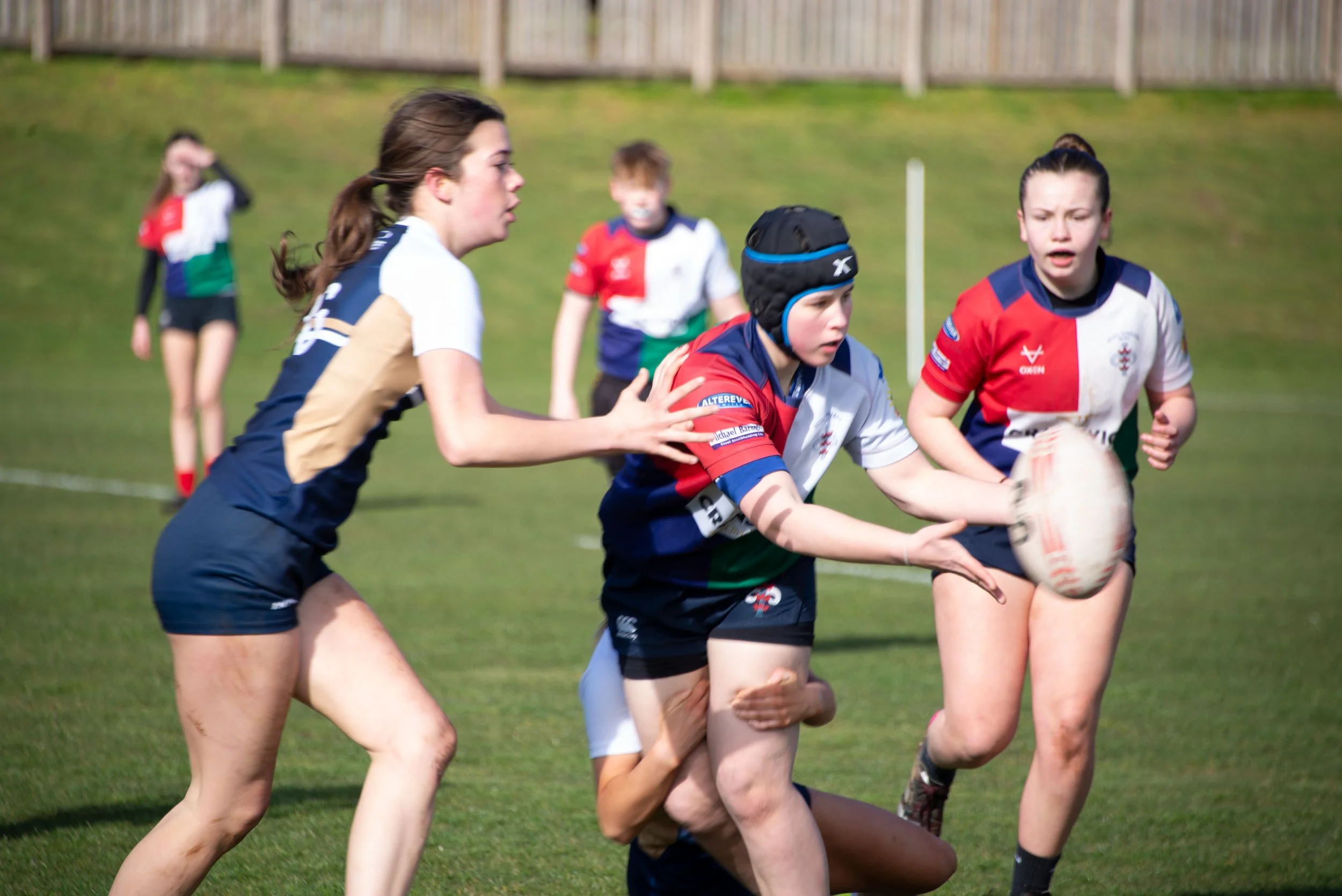 Girls playing rugby on a grassy field during daytime, some wearing protective headgear.