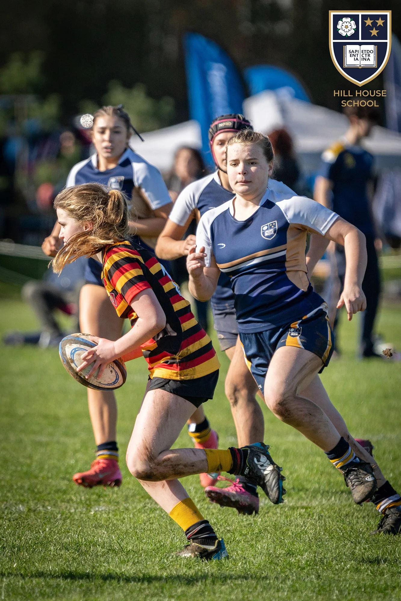 Young girls playing rugby on a grassy field during daytime, wearing sports uniforms. One girl with long hair in a red and yellow striped jersey holds a rugby ball, while others in blue and white jerseys run behind her. There are tents and flags in th