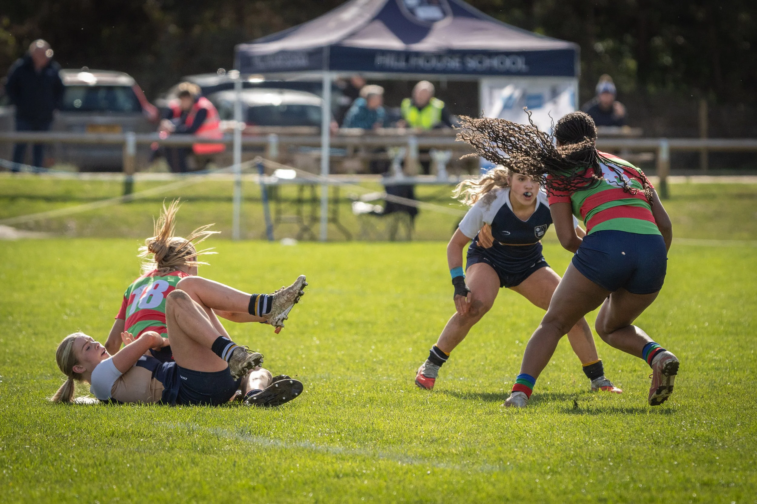 Rugby match with female players, some on the ground and others in motion on green grass field, with spectators and a tent in the background.