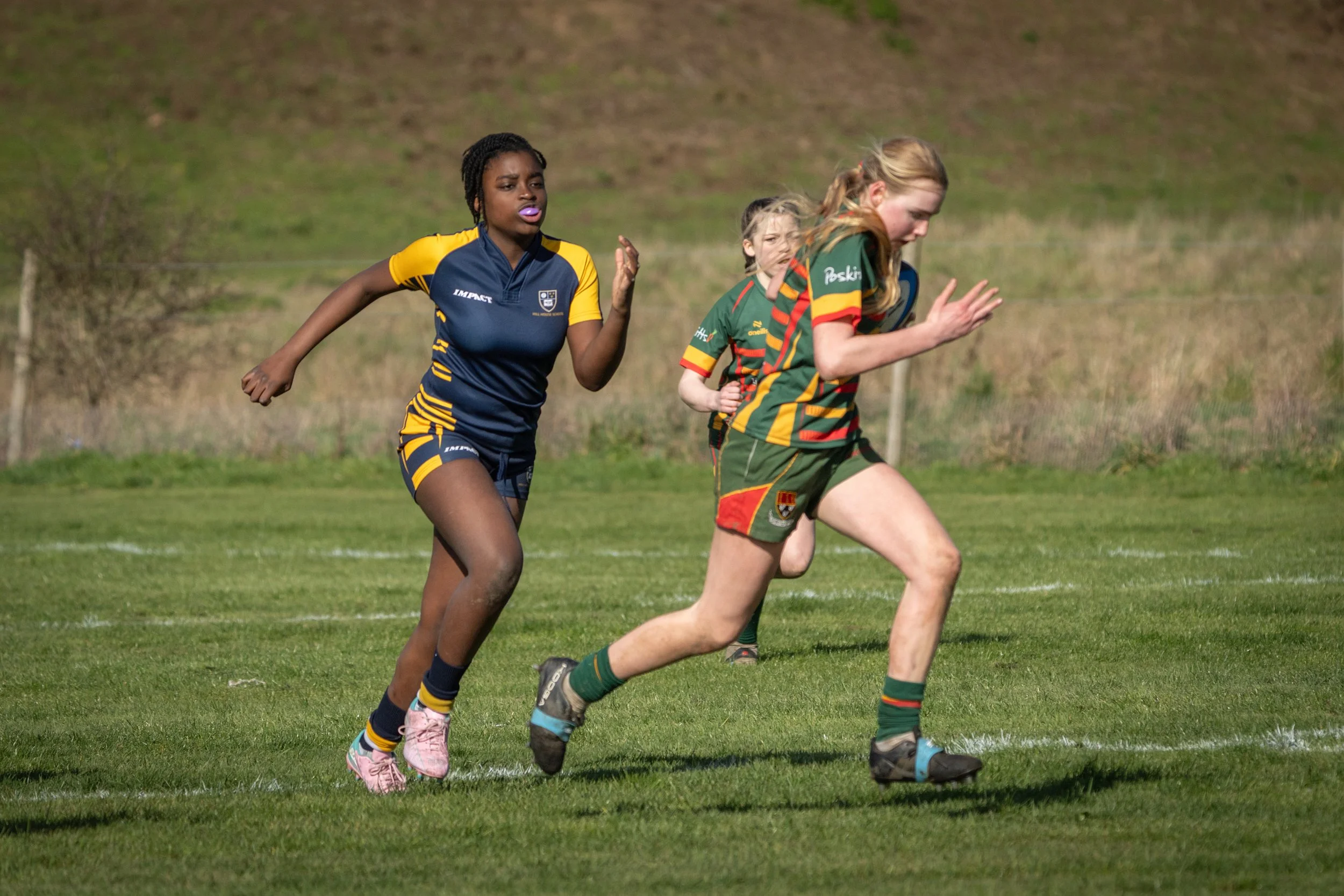 Women and girls running on a grassy field during a sports activity, wearing colorful sports uniforms.