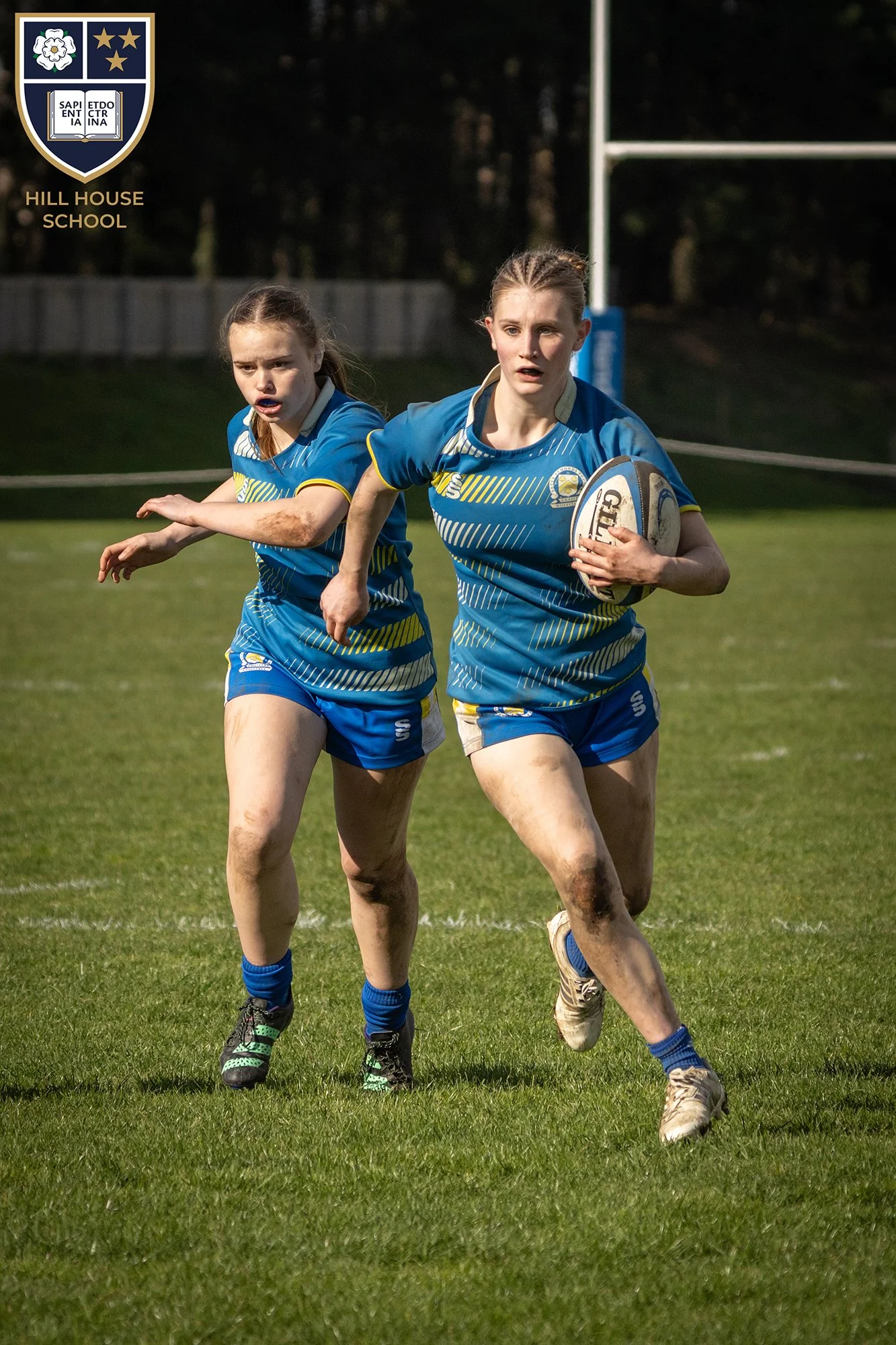 Two young female rugby players running on a grass field, wearing blue and yellow uniforms, with muddy legs and feet, one holding a rugby ball.
