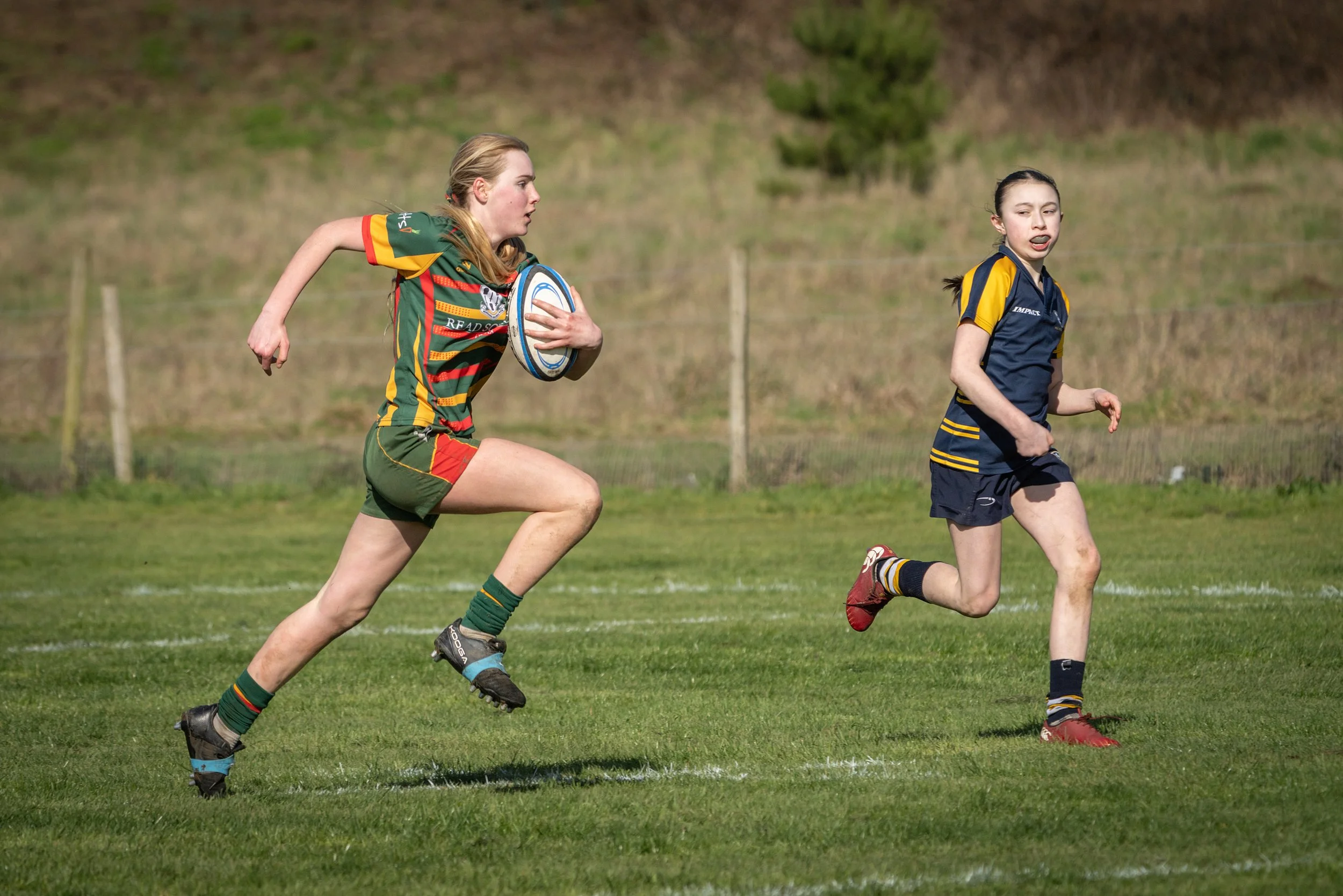 Two young female rugby players running on a grassy field during a game, one with a rugby ball, in colorful uniforms and athletic gear.