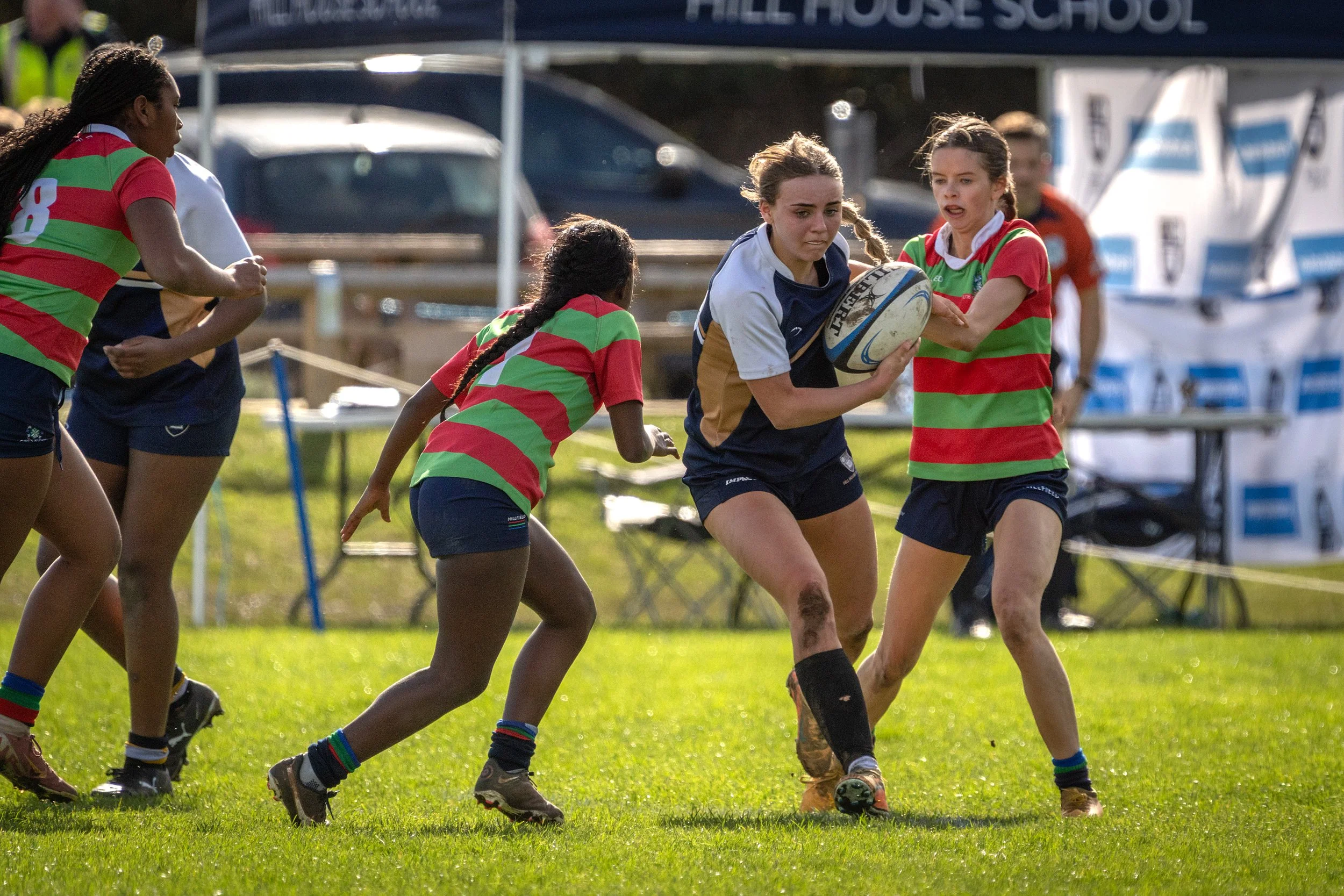 Women playing rugby on a grassy field, with some wearing red and green striped jerseys, and one woman holding a rugby ball while others attempt to block or tackle her.