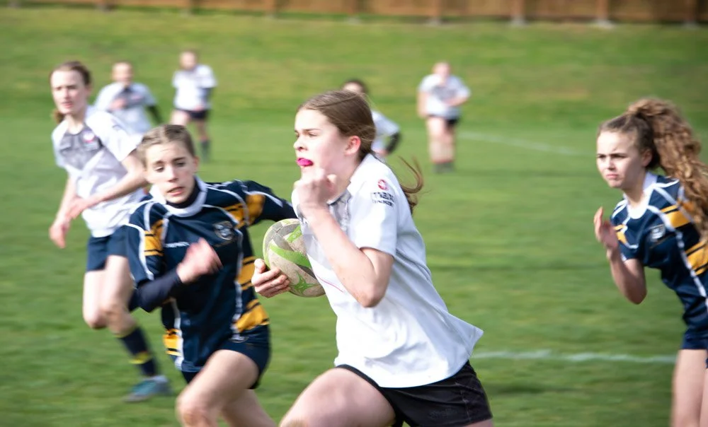 Young girls playing rugby on a grassy field, with some running in the background.
