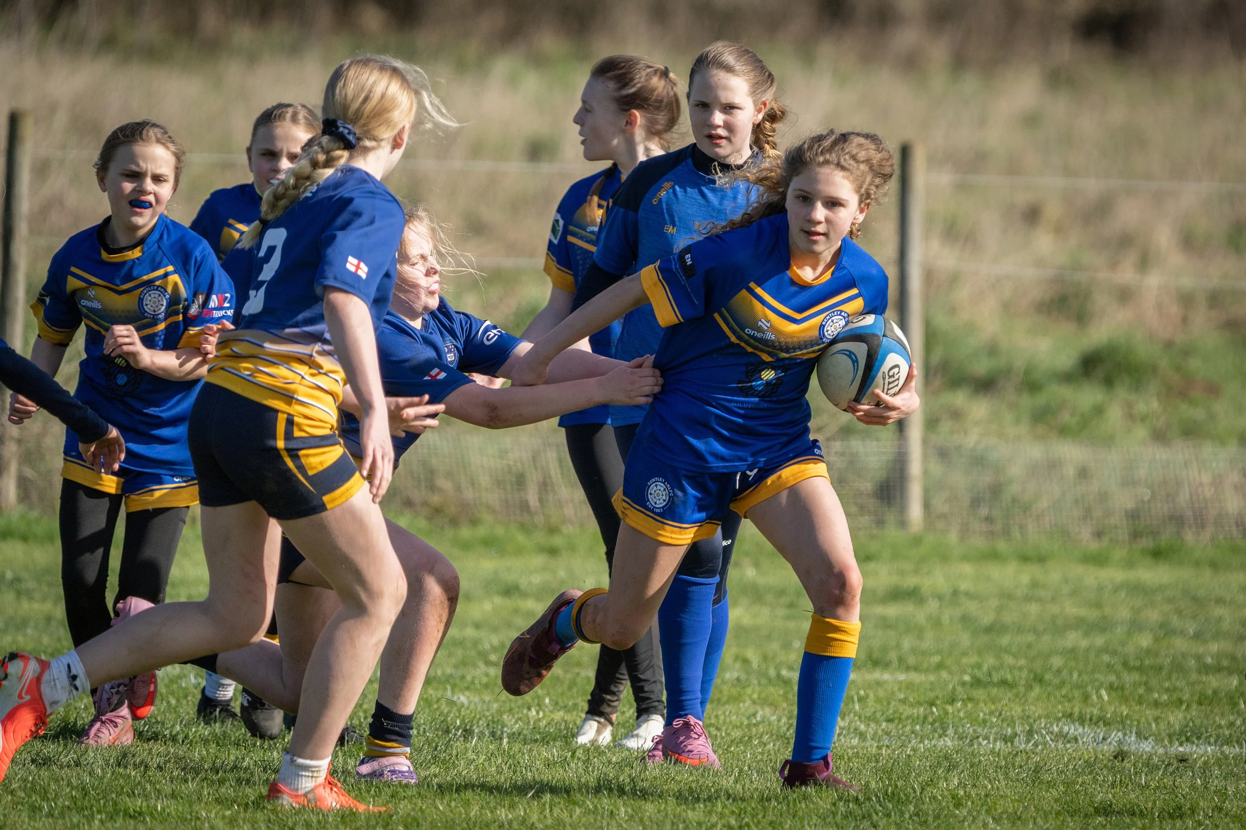 Young girls playing rugby on a grassy field, wearing blue sports uniforms, some with braided hair, engaging in a competitive game with one girl holding a rugby ball.