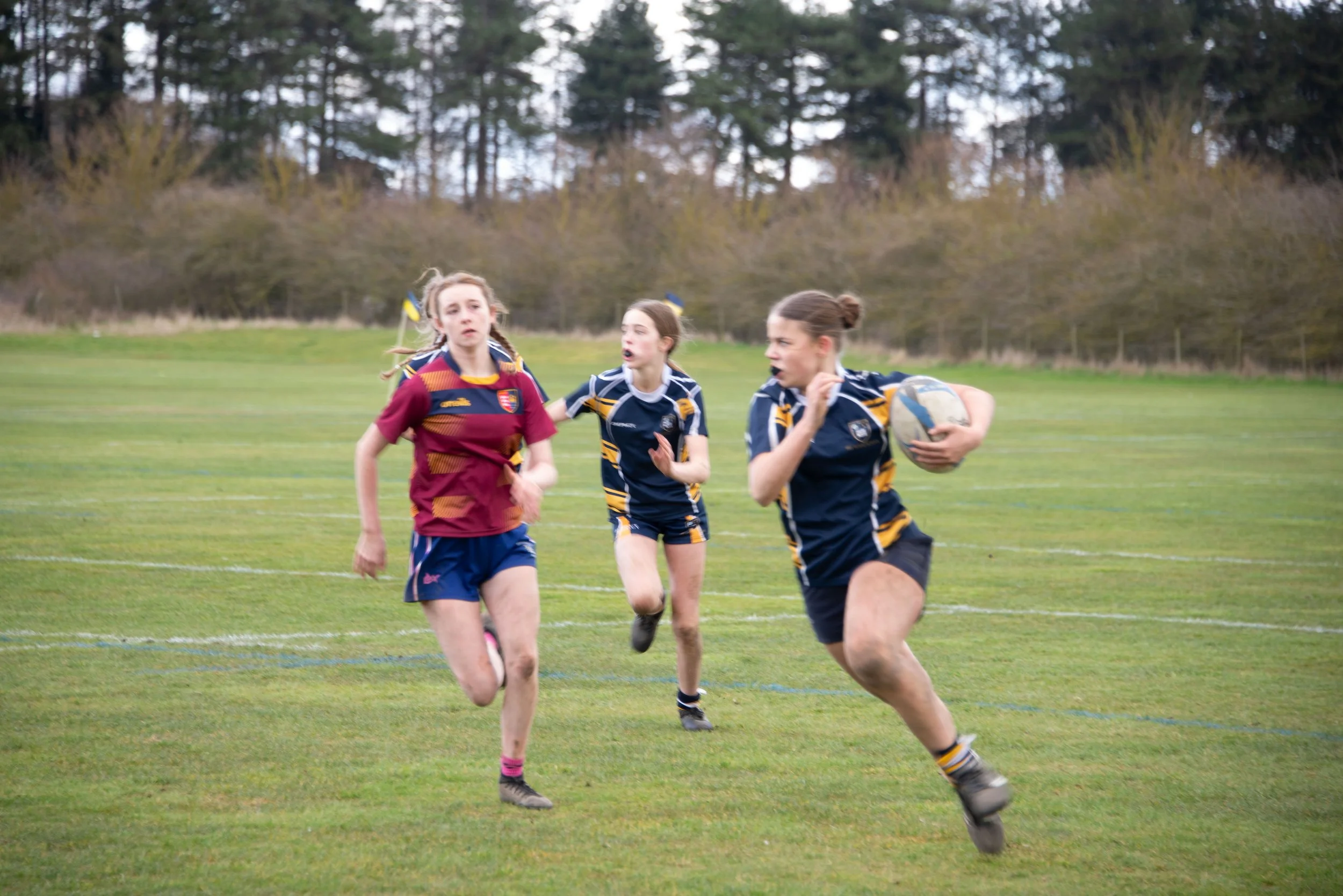 Young girls playing rugby on a grassy field with trees in the background, wearing sports uniforms and running with a rugby ball.