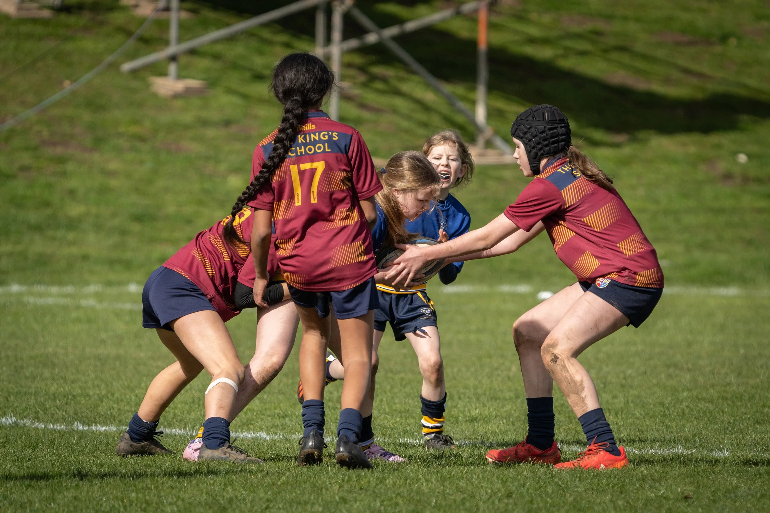 Girls playing rugby on the field, wearing team jerseys, with one holding the ball and others reaching for it.