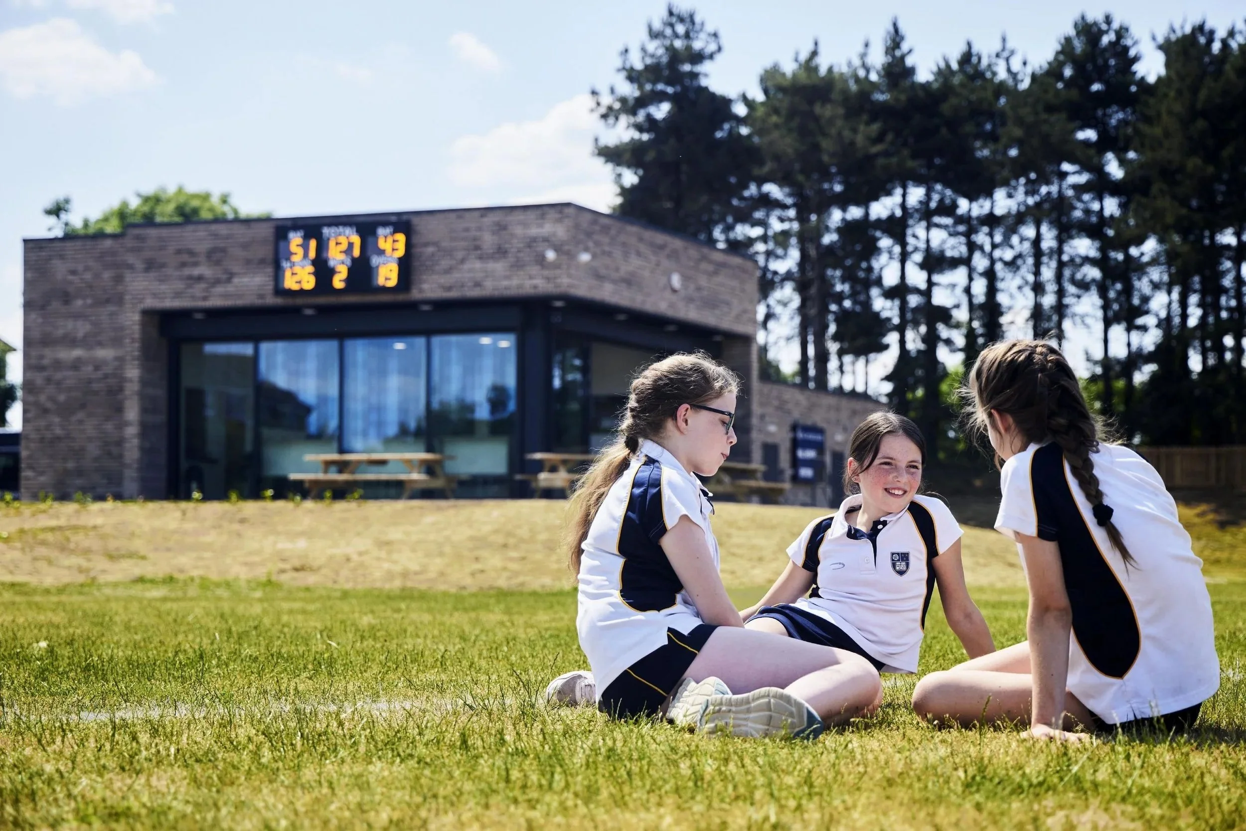 Four young girls in school uniforms sitting and talking on a grassy field with a modern brick building and digital scoreboard in the background.