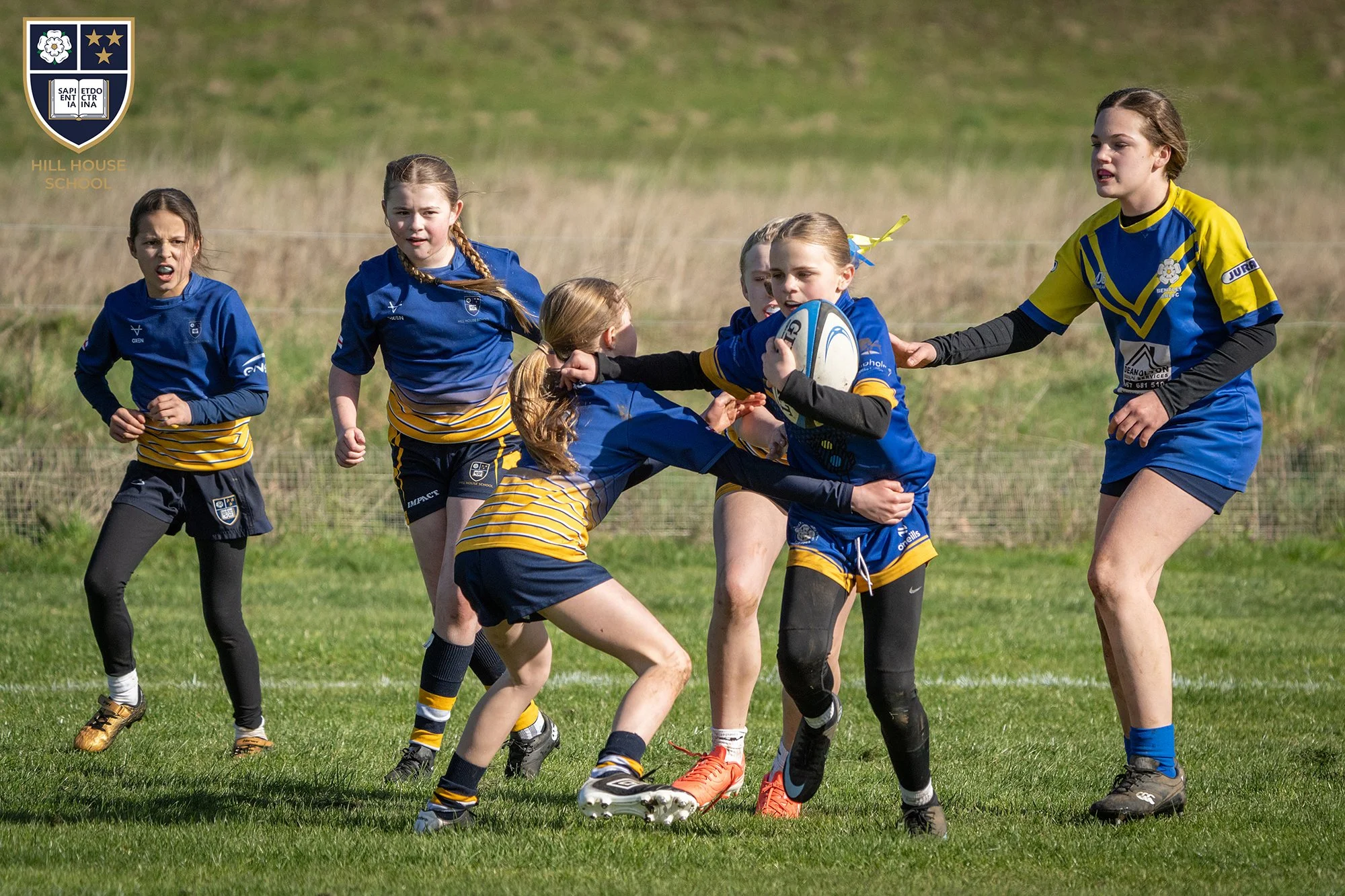 Girls playing rugby on a grassy field, with some girls wearing blue and yellow uniforms and others in dark sportswear. One girl is holding a rugby ball, and there is a girl in a yellow and blue jersey reaching out to her. The background shows a grass